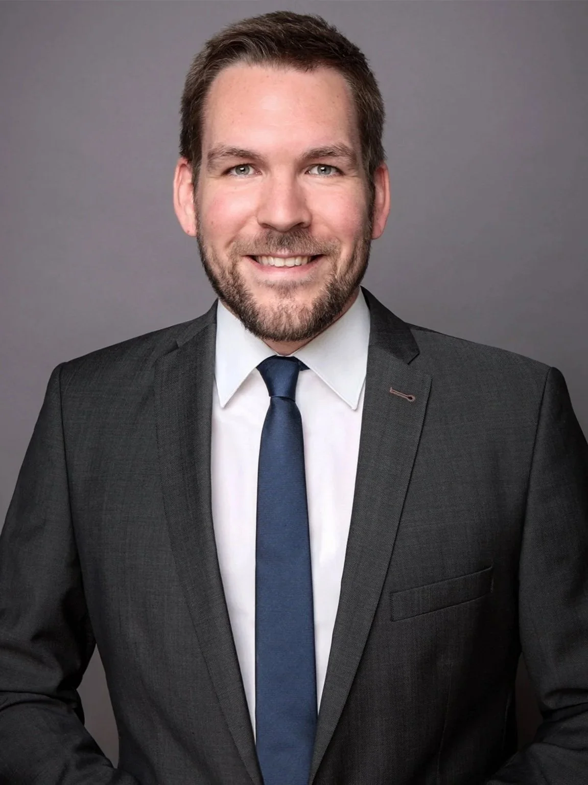 Ein Mann in einem dunklen Anzug, weißem Hemd und blauer Krawatte vor einem grauen Hintergrund.