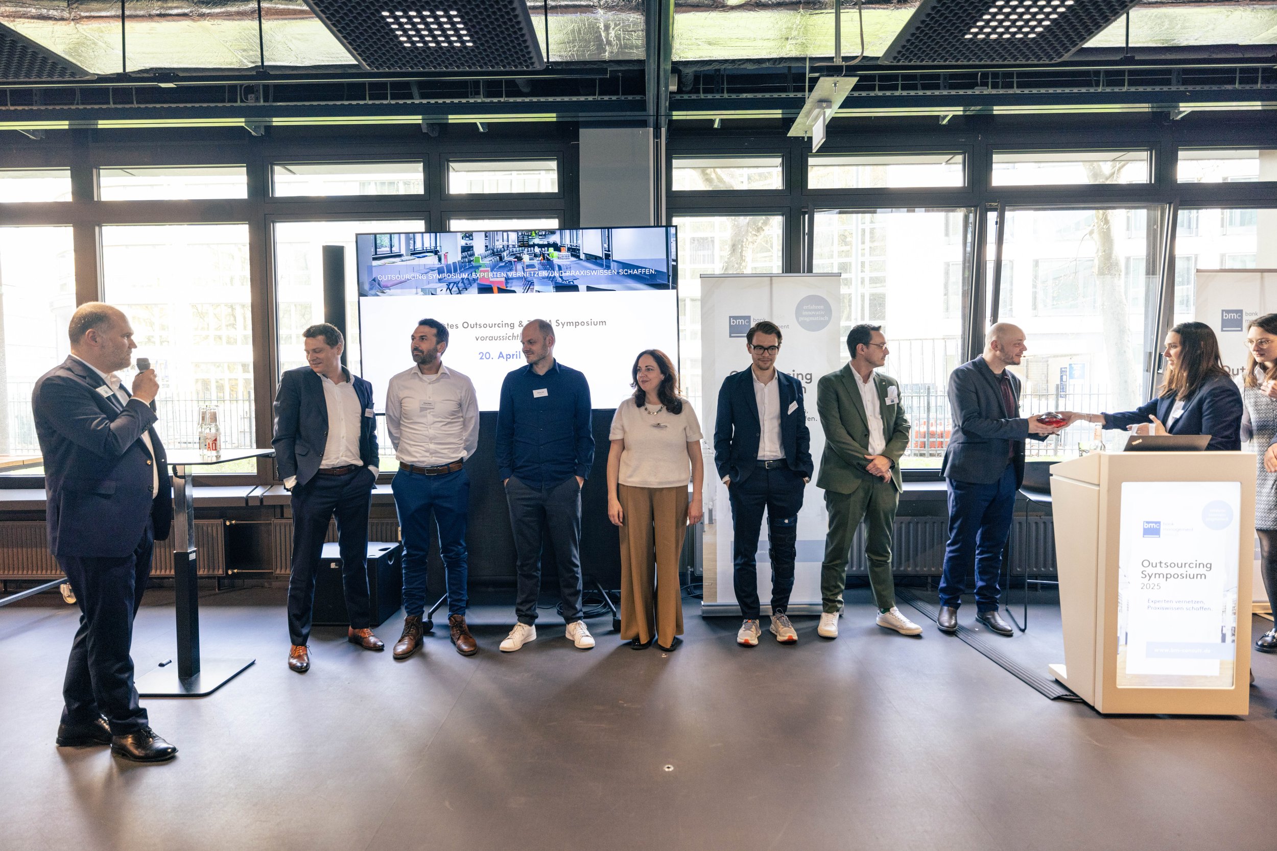 Eine Gruppe von neun Personen bei einem OutSourcing-Symposium im Raum, einige auf der Bühne, eine Frau erhält eine Auszeichnung. Es gibt einen großen Bildschirm und ein Podium.