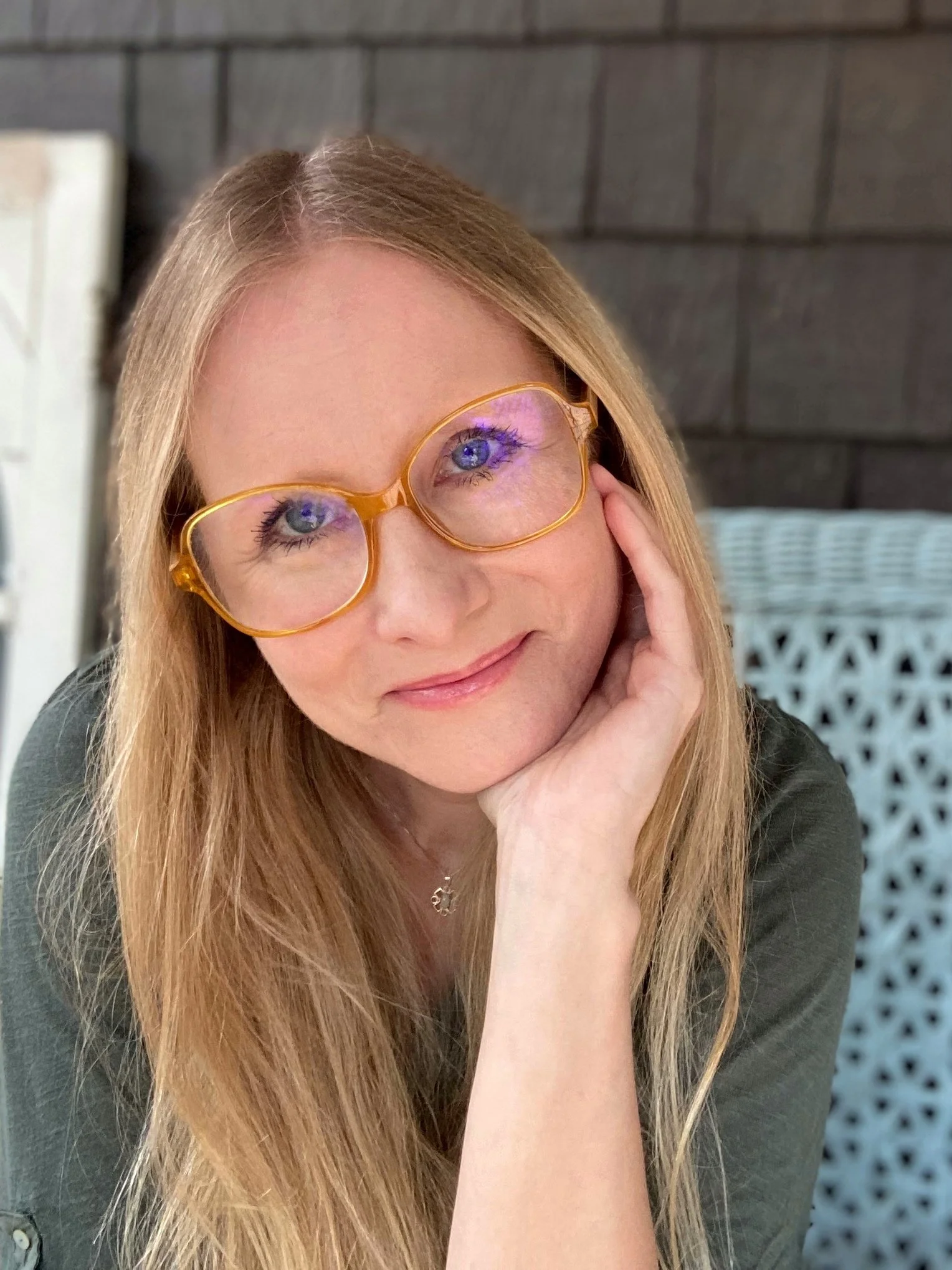 A woman with long blonde hair, wearing large, round, orange-rimmed glasses, smiling softly while resting her head on her hand, sitting outdoors near a dark wooden wall.