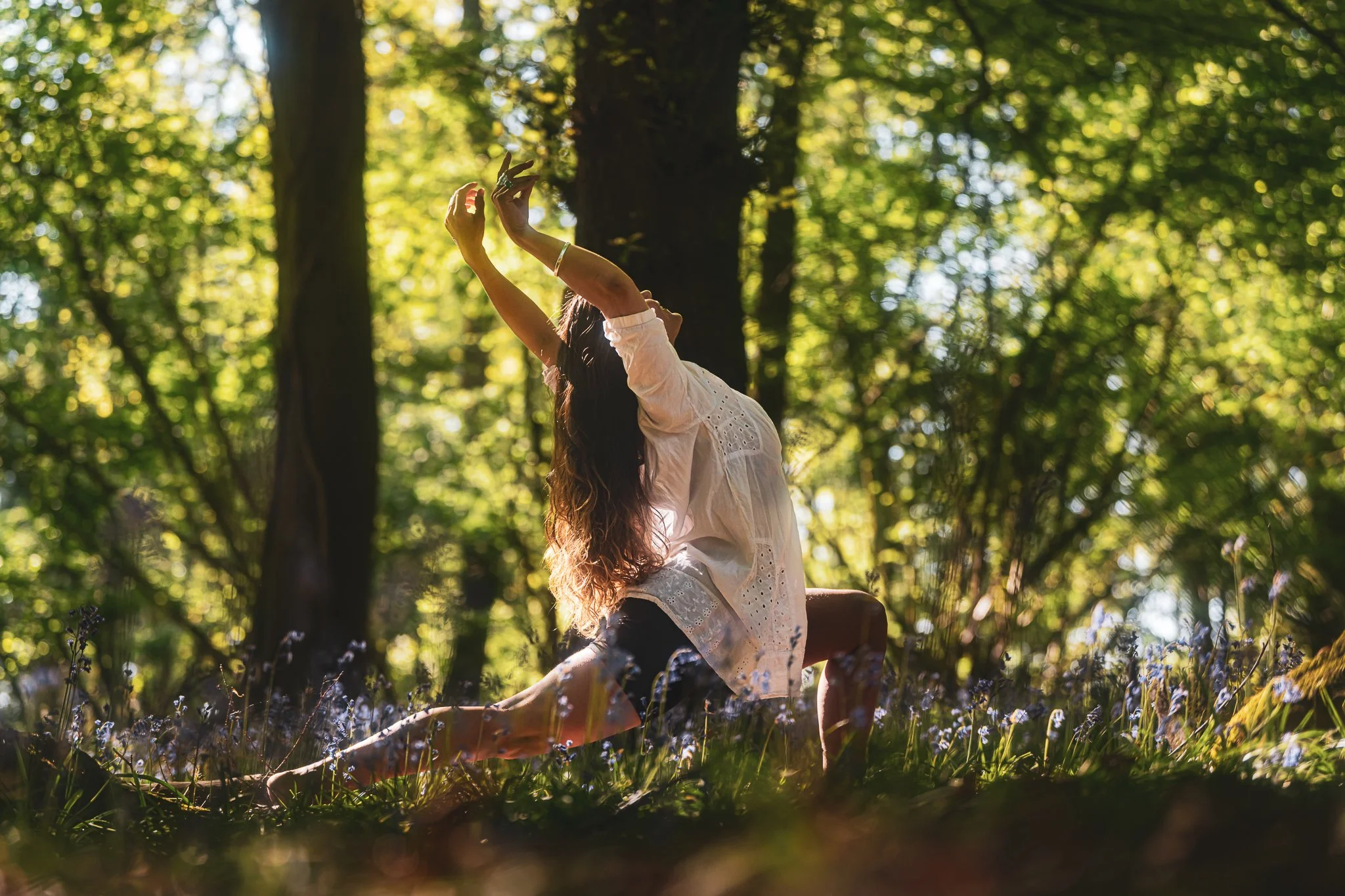 A woman practicing yoga outdoors in a lush forest, performing a lunge pose with arms raised overhead on a wooden bench surrounded by greenery and sunlight filtering through trees.