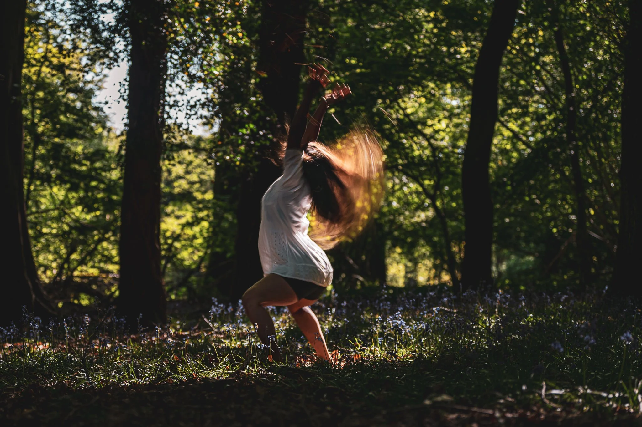 A woman with long hair, wearing a white dress and black shorts, dancing or spinning in a forest with sunlight filtering through the trees, creating a dreamy atmosphere.