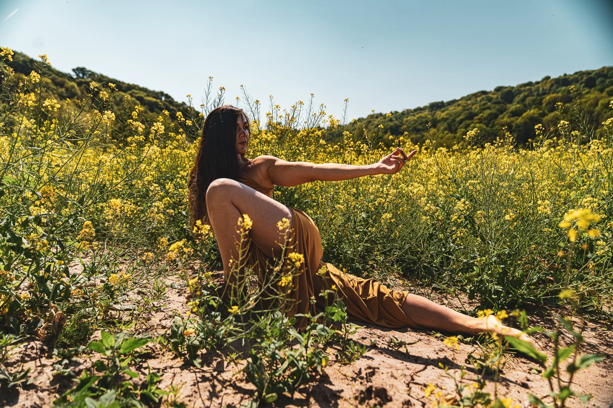 A woman with long dark hair sitting among yellow wildflowers in a field, reaching out with her right arm, with green hills in the background on a sunny day.