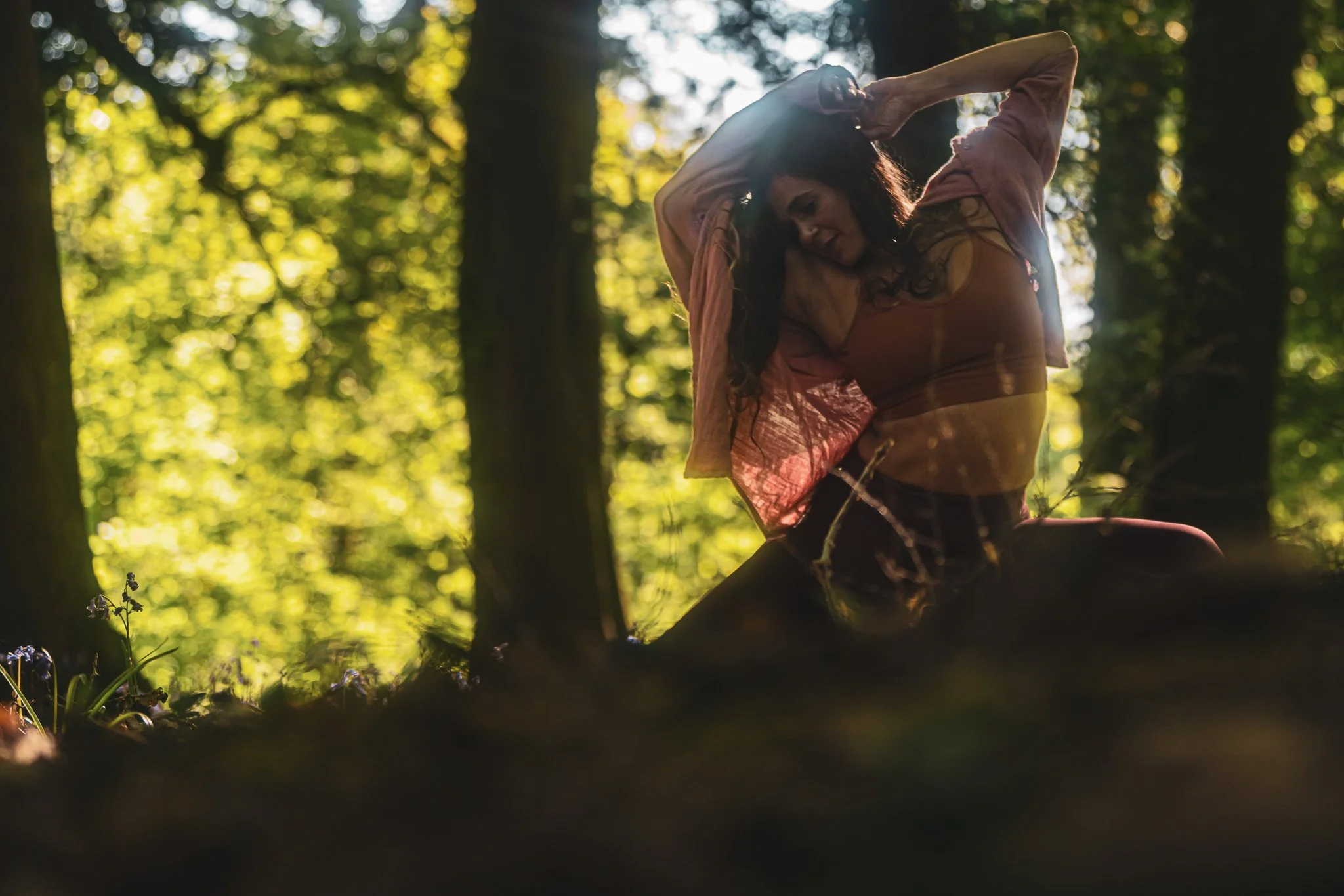 A woman with dark hair, wearing a sheer top and pink jacket, is sitting on the ground in a forest with sunlight filtering through the trees.