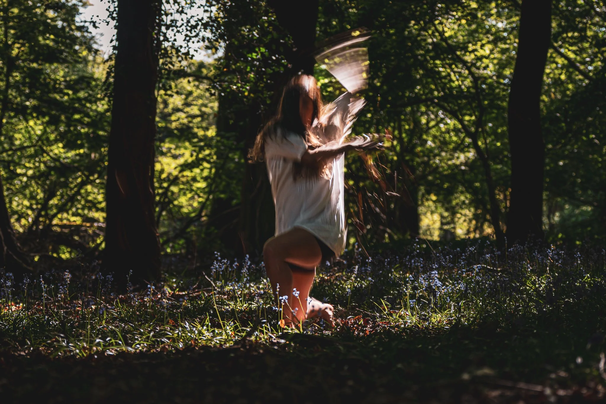 A woman in a forest surrounded by tall trees and small blue flowers, spinning and tossing her hair.