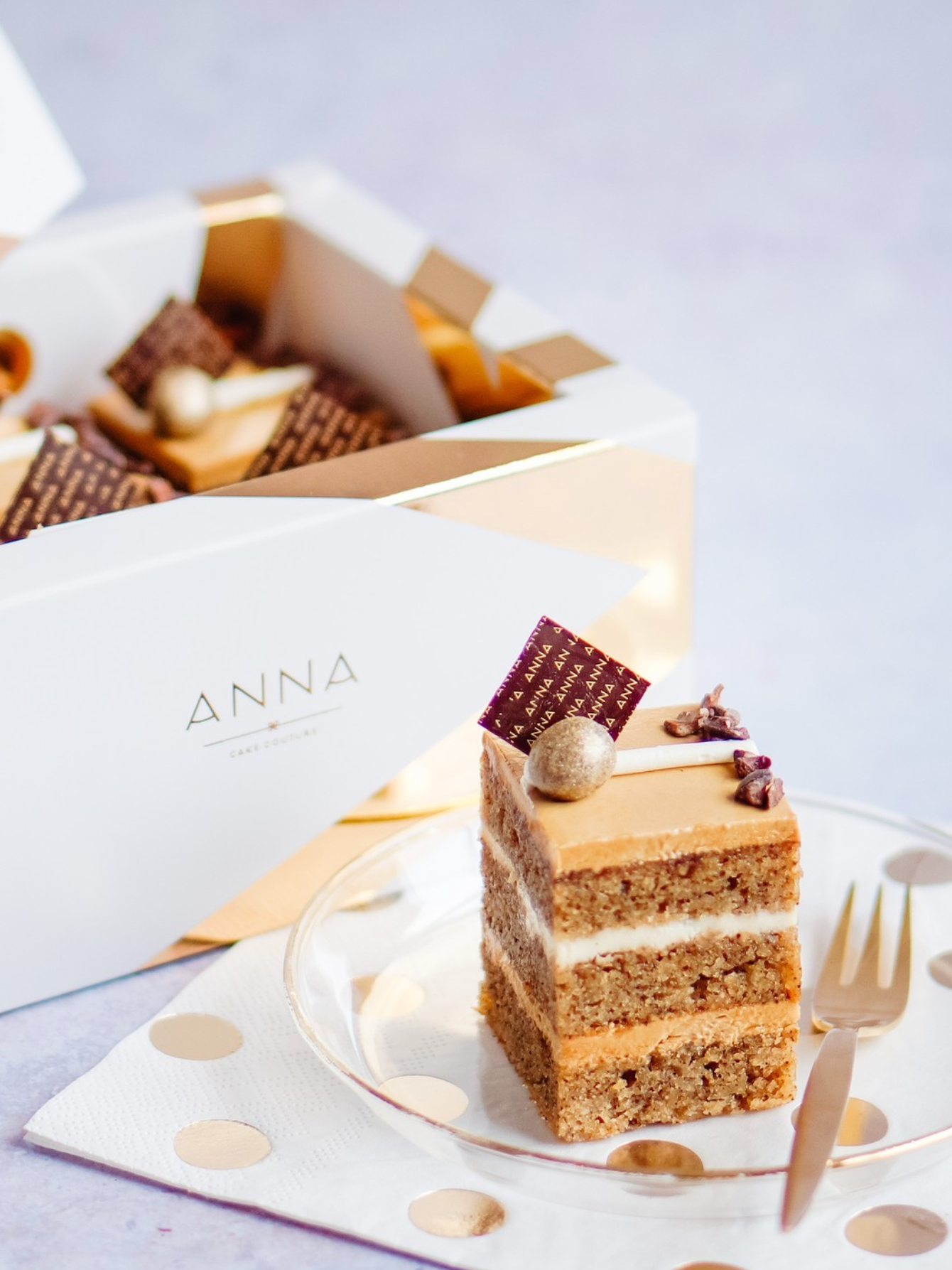 A slice of layered cake with caramel-colored frosting on a clear plate, decorated with a small round edible metallic decoration and a rectangular chocolate piece with gold writing, near a fork. In the background, there are boxes of assorted chocolate