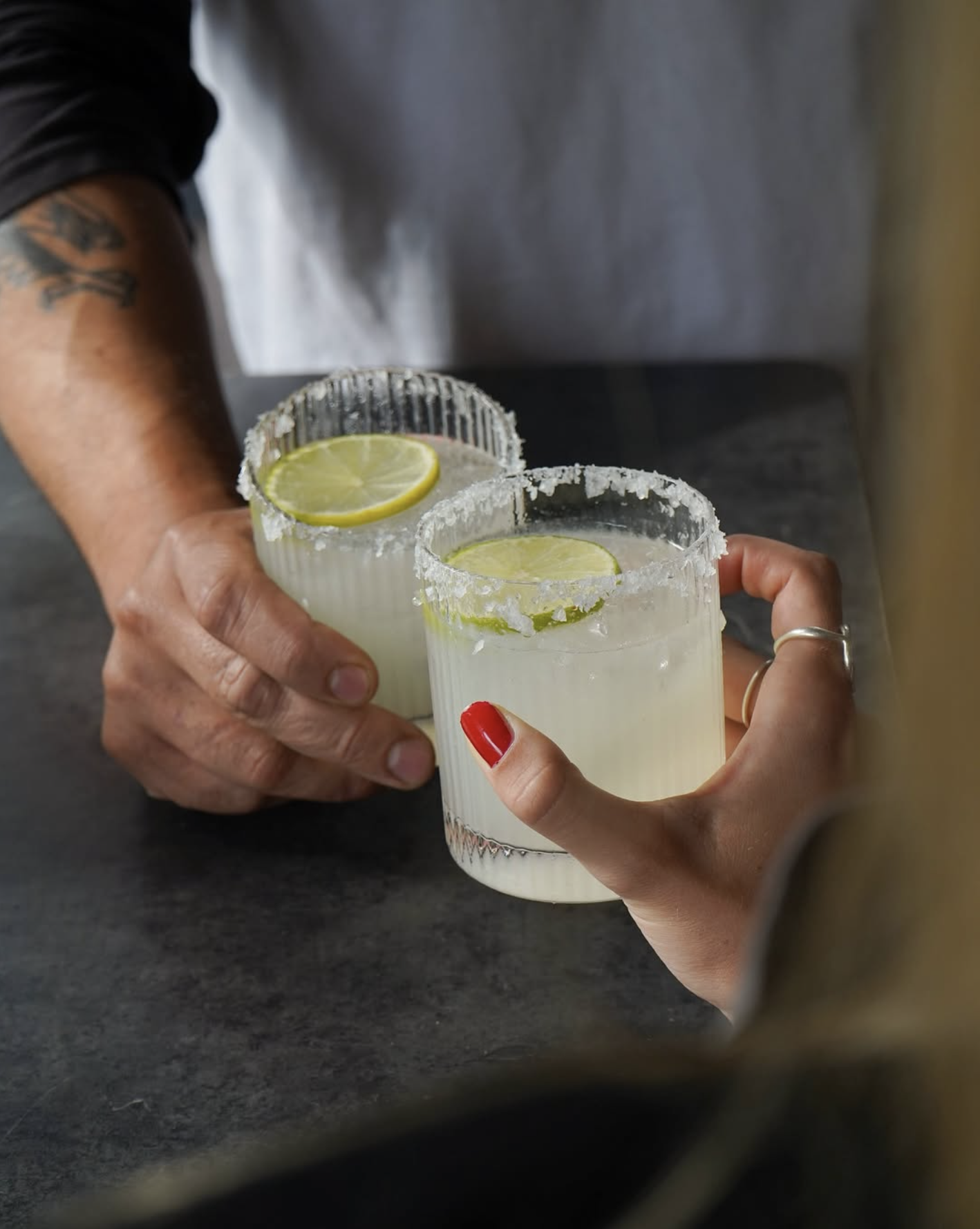 Two margarita glasses with salted rims and lime slices on top, being held by two people, one with red nail polish, on a dark table.