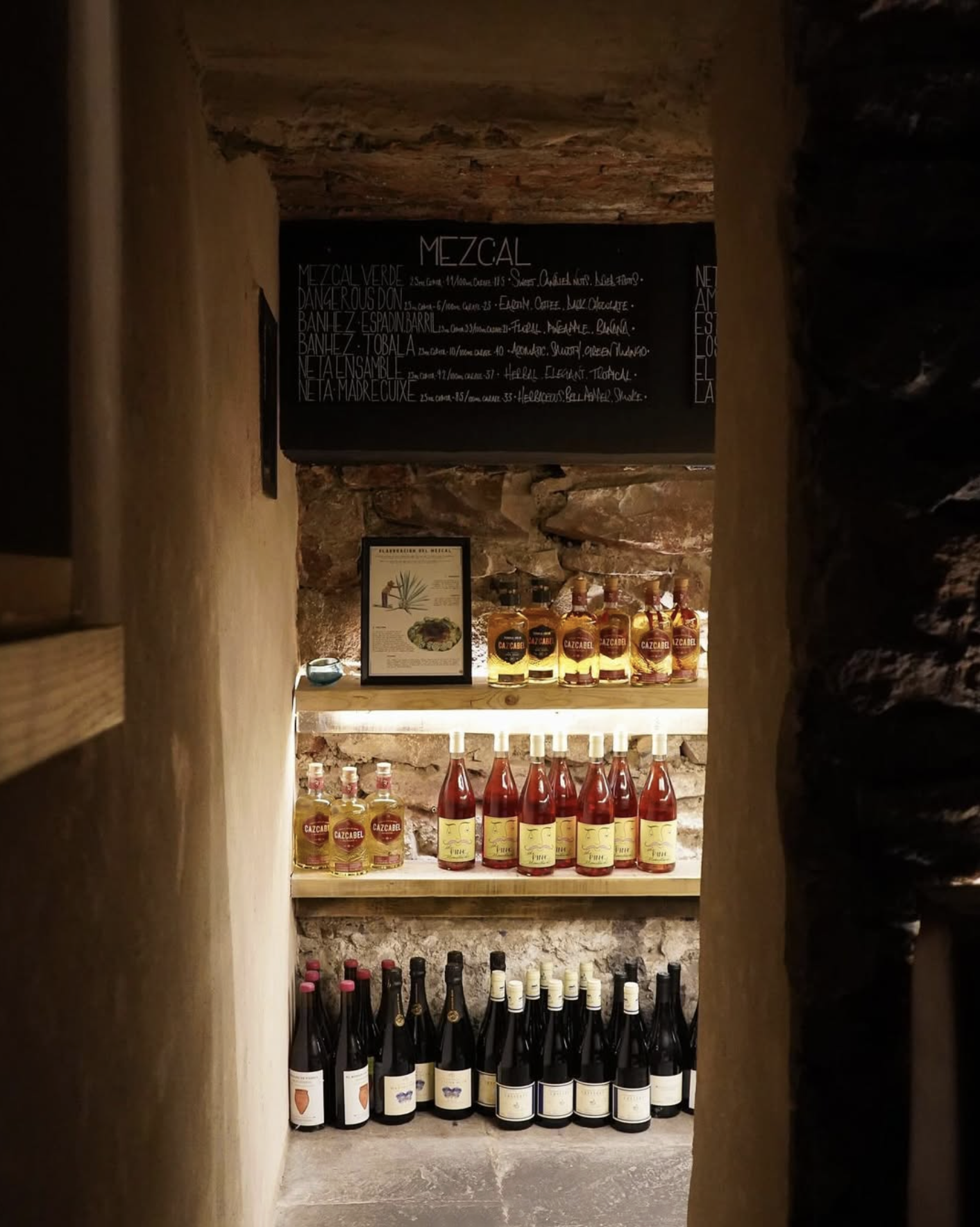 Shelves with bottles of liquor and wine, framed picture, chalkboard menu, in a rustic cellar or bar setting.