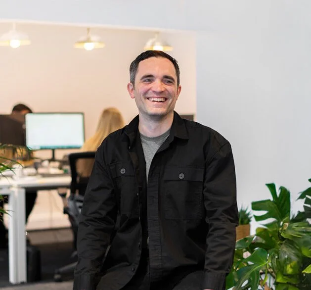 A smiling man in a black jacket sitting in an office with coworkers and plants in the background.