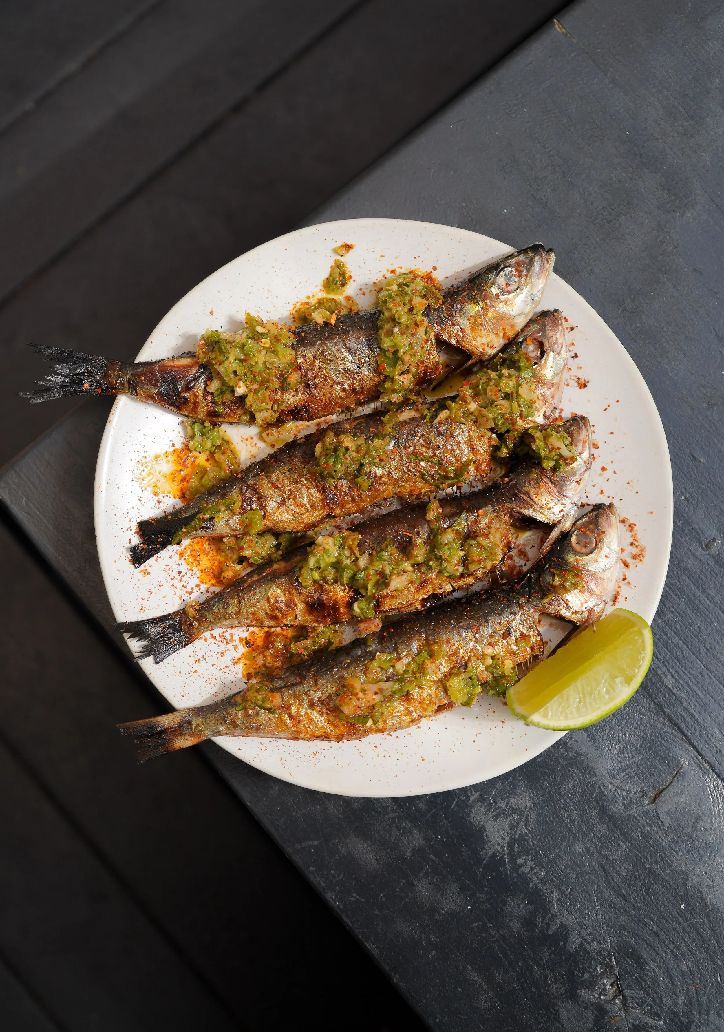 Plate of grilled sardines garnished with chopped green herbs and a lime wedge.