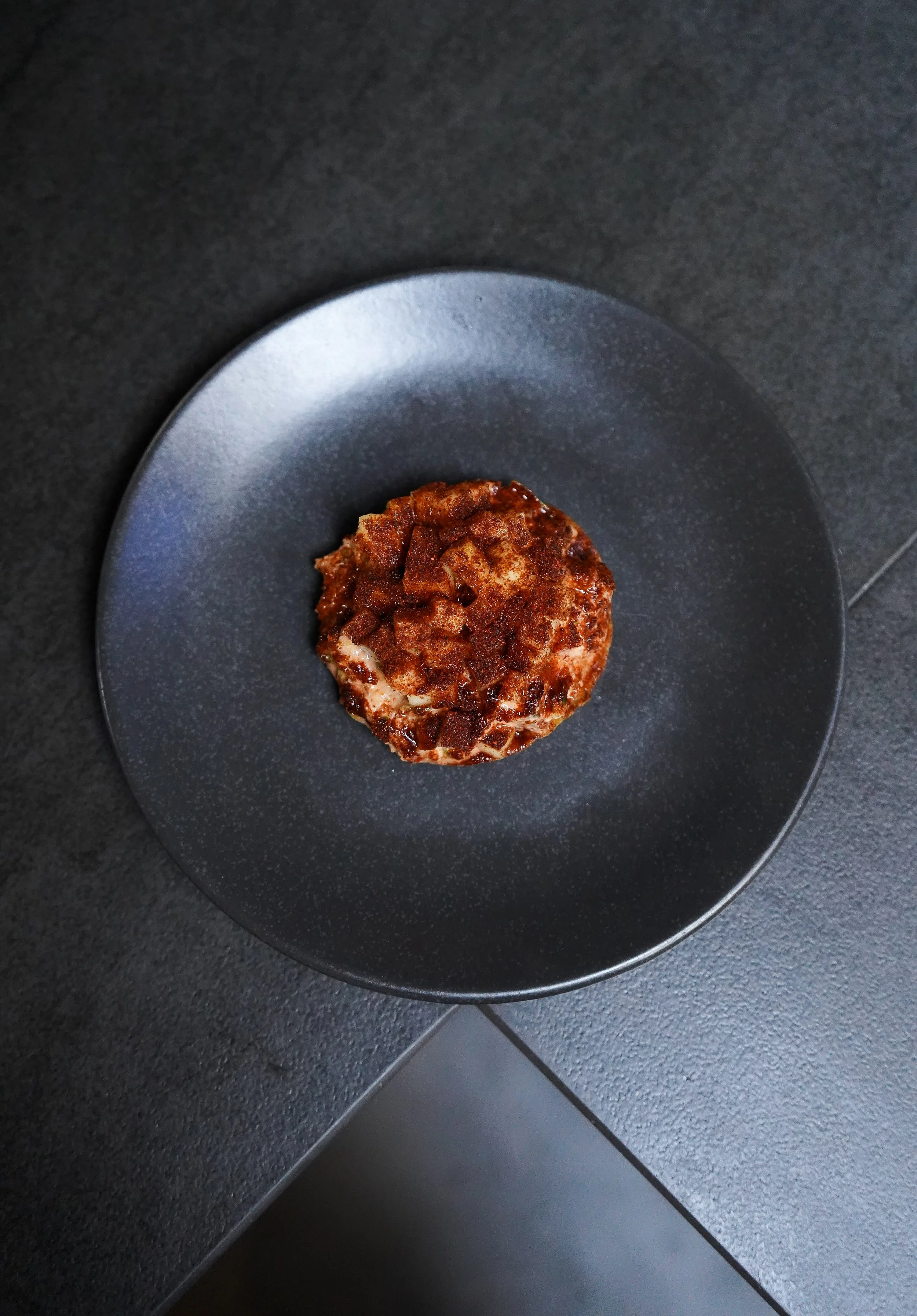 A chocolate dessert topped with chocolate chunks on a dark plate.