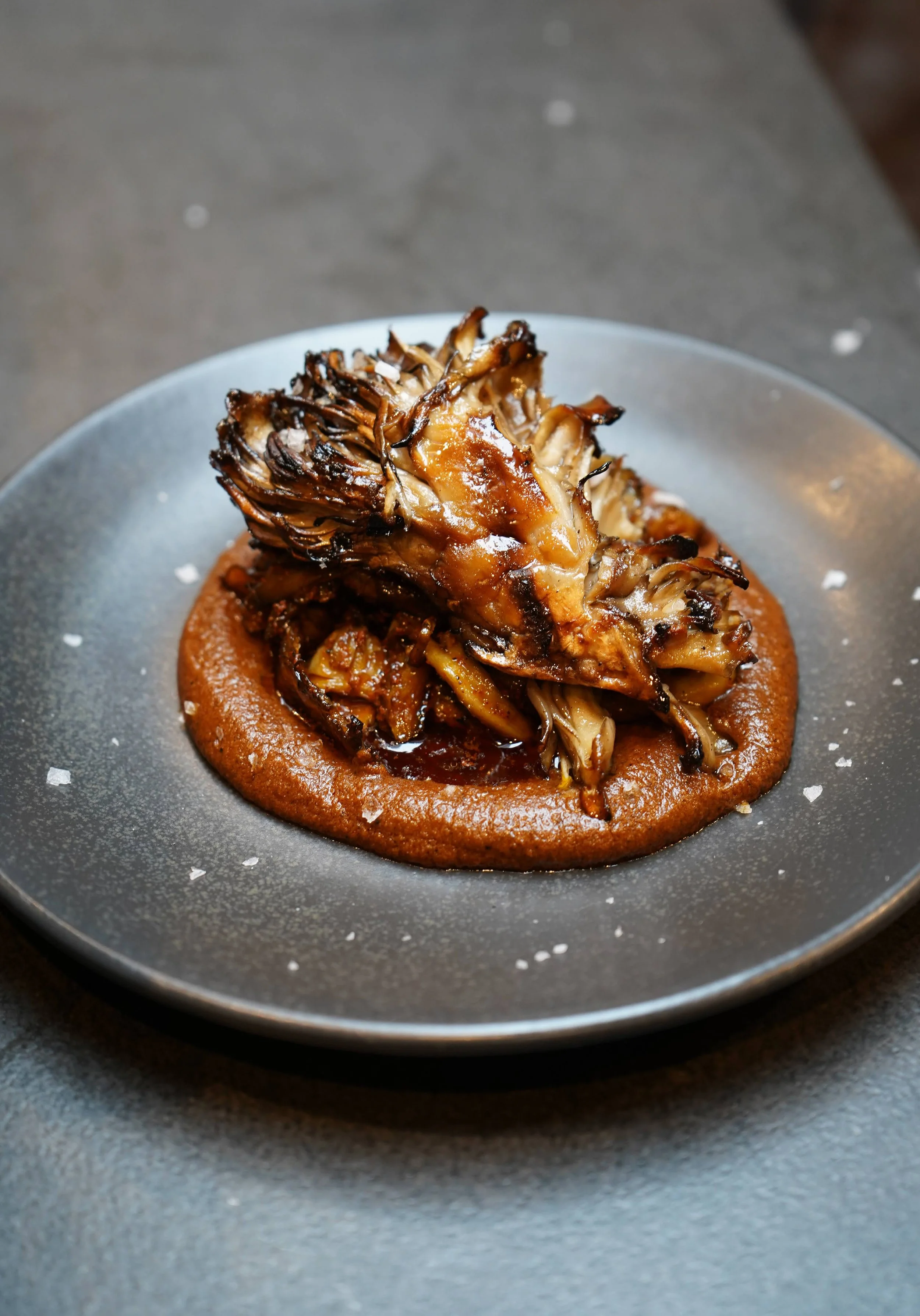 Roasted artichoke atop a smear of red sauce on a dark gray plate, garnished with sea salt.