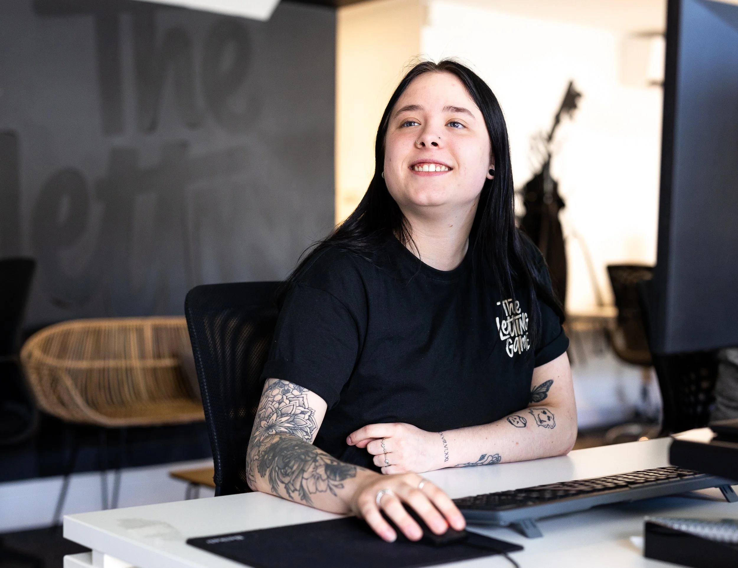 A young woman with black hair and tattoos sitting at a desk in an office, smiling and looking slightly to the side.