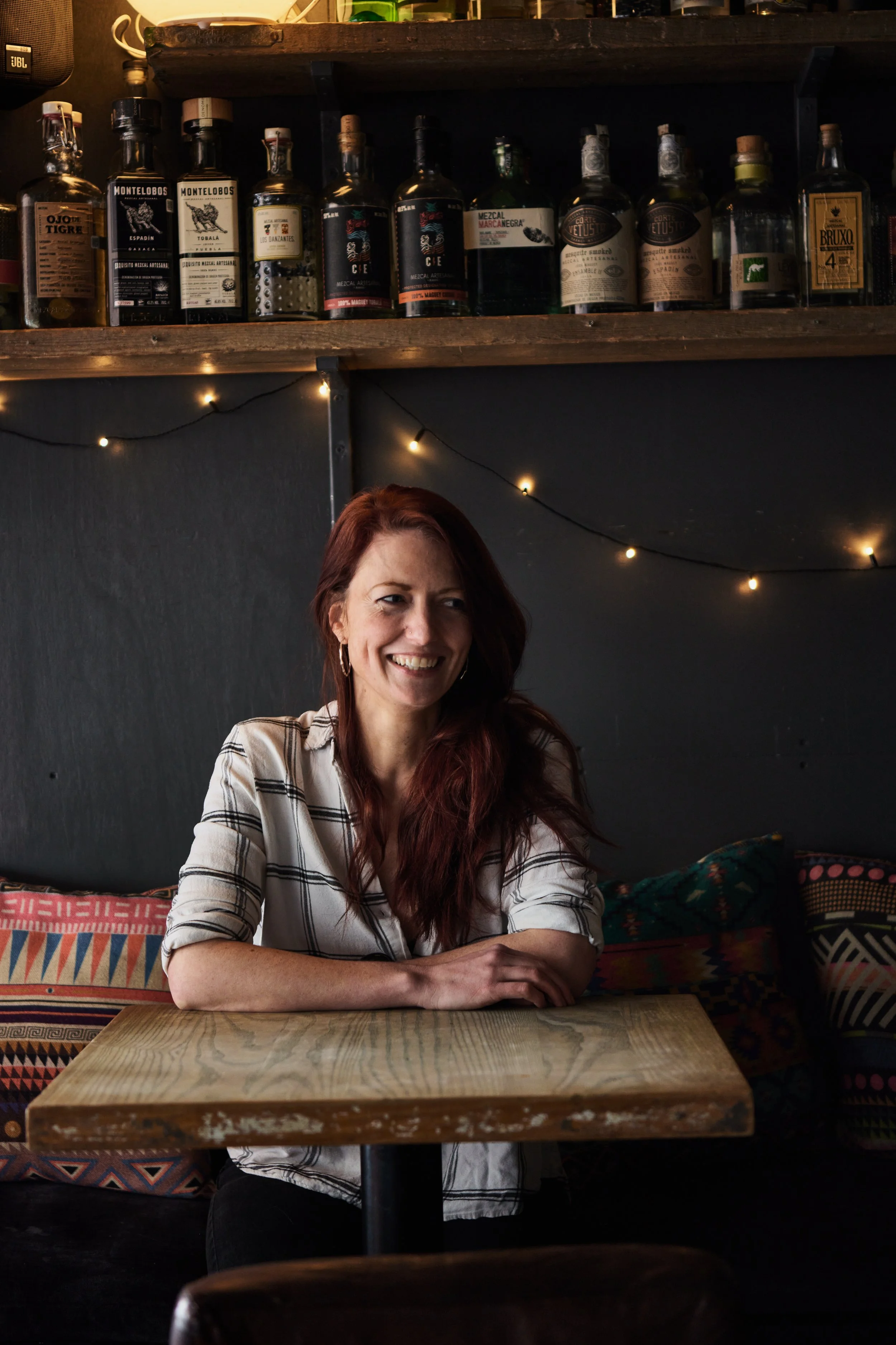 A woman with long red hair smiling while sitting at a wooden table in a dimly lit room with dark walls, decorated with colorful pillows and string lights. Behind her is a shelf with various bottles of alcohol.