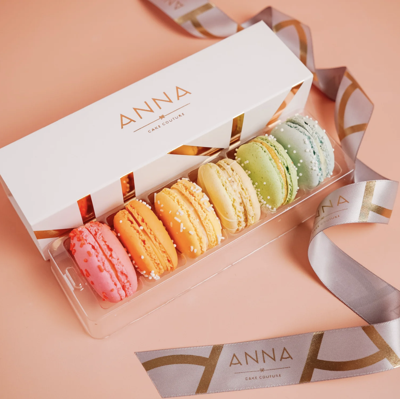 A white box labeled 'Anna Cake Couture' contains six colorful macarons with different pastel shades, decorated with sugar pearls, placed on a pink surface with a silver and gold ribbon nearby.