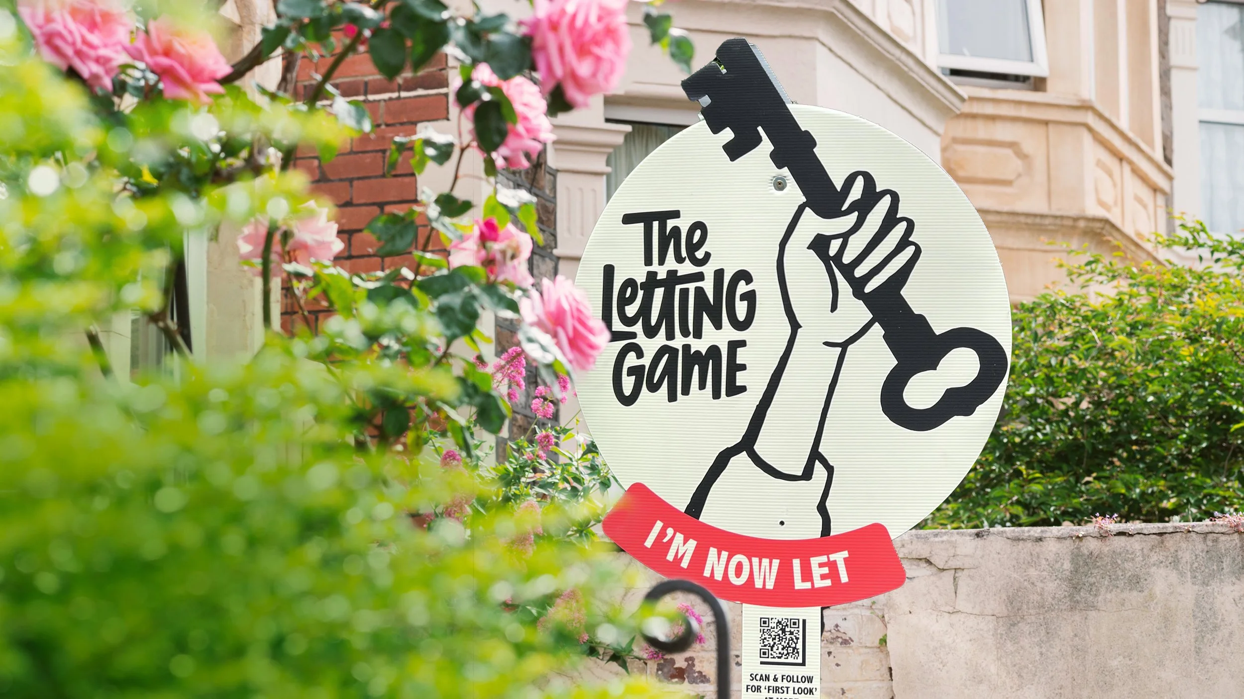 Sign with a hand holding a key, with text that says "The Letting Game" and "I'm now let." The sign is surrounded by pink flowers and green foliage, with residential buildings in the background.