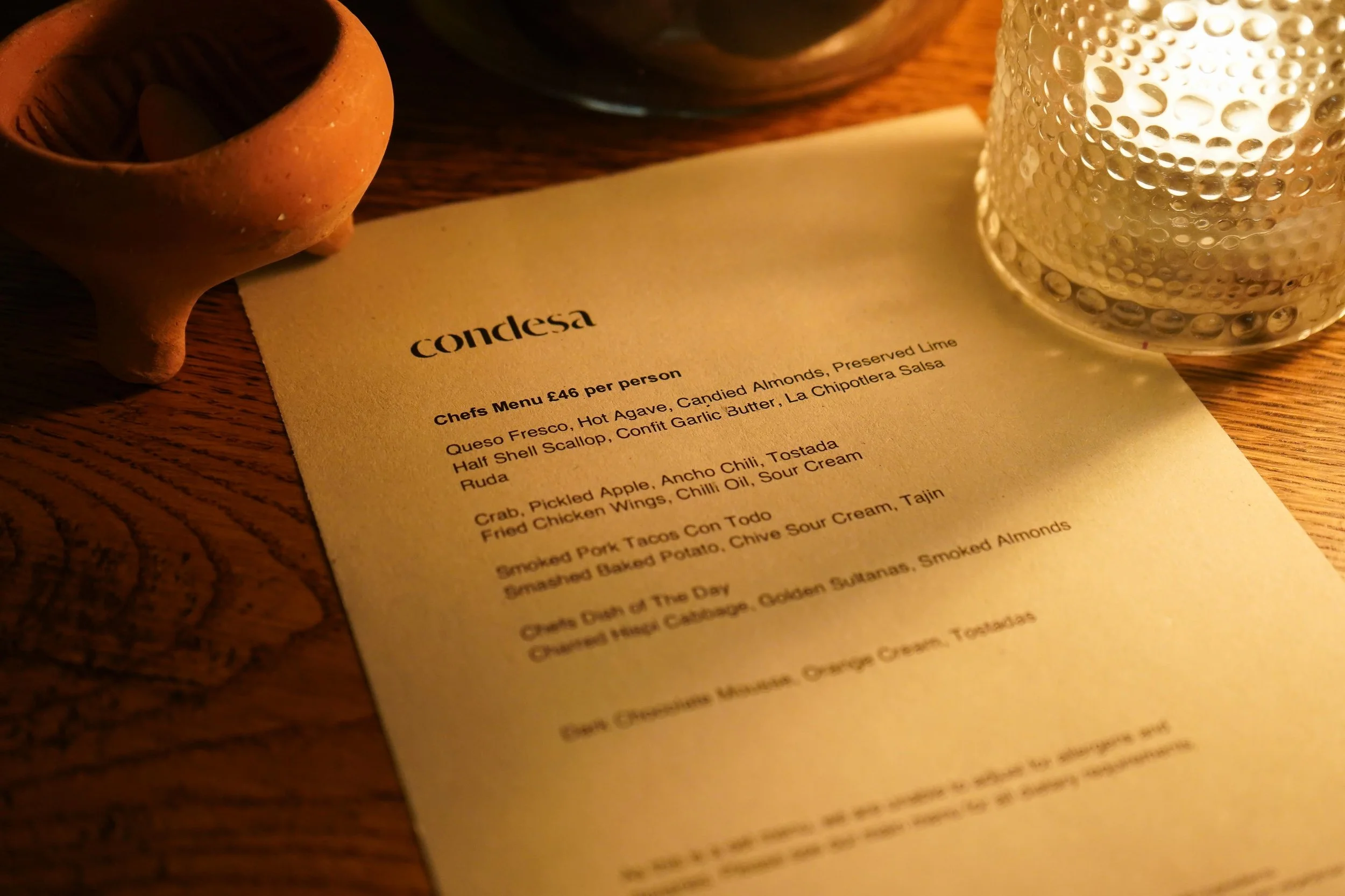 A restaurant menu from Condesa, featuring various dishes, on a wooden table with a ceramic vessel, a glass candle holder, and a bowl visible nearby.