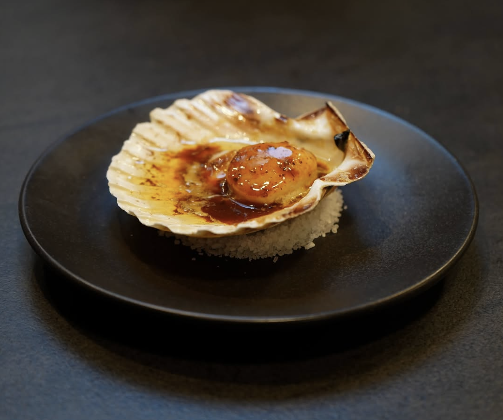 A gourmet dish featuring a baked scallop in its shell topped with a poached egg, sauce, and garnished with a bed of seasoned rice on a black plate.