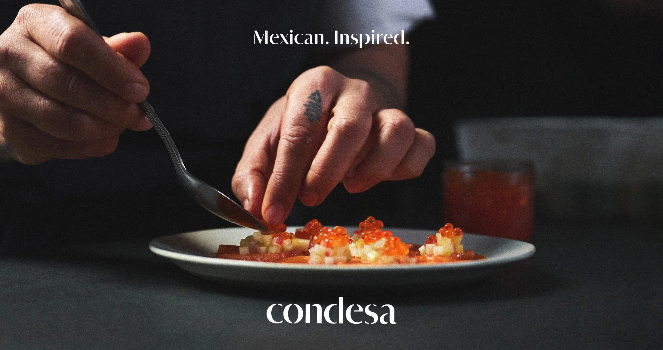 Close-up of a person’s hands preparing Mexican-inspired food on a white plate, with a dark background and a small glass of red sauce nearby, along with the text "Mexican. Inspired." and "condesa".