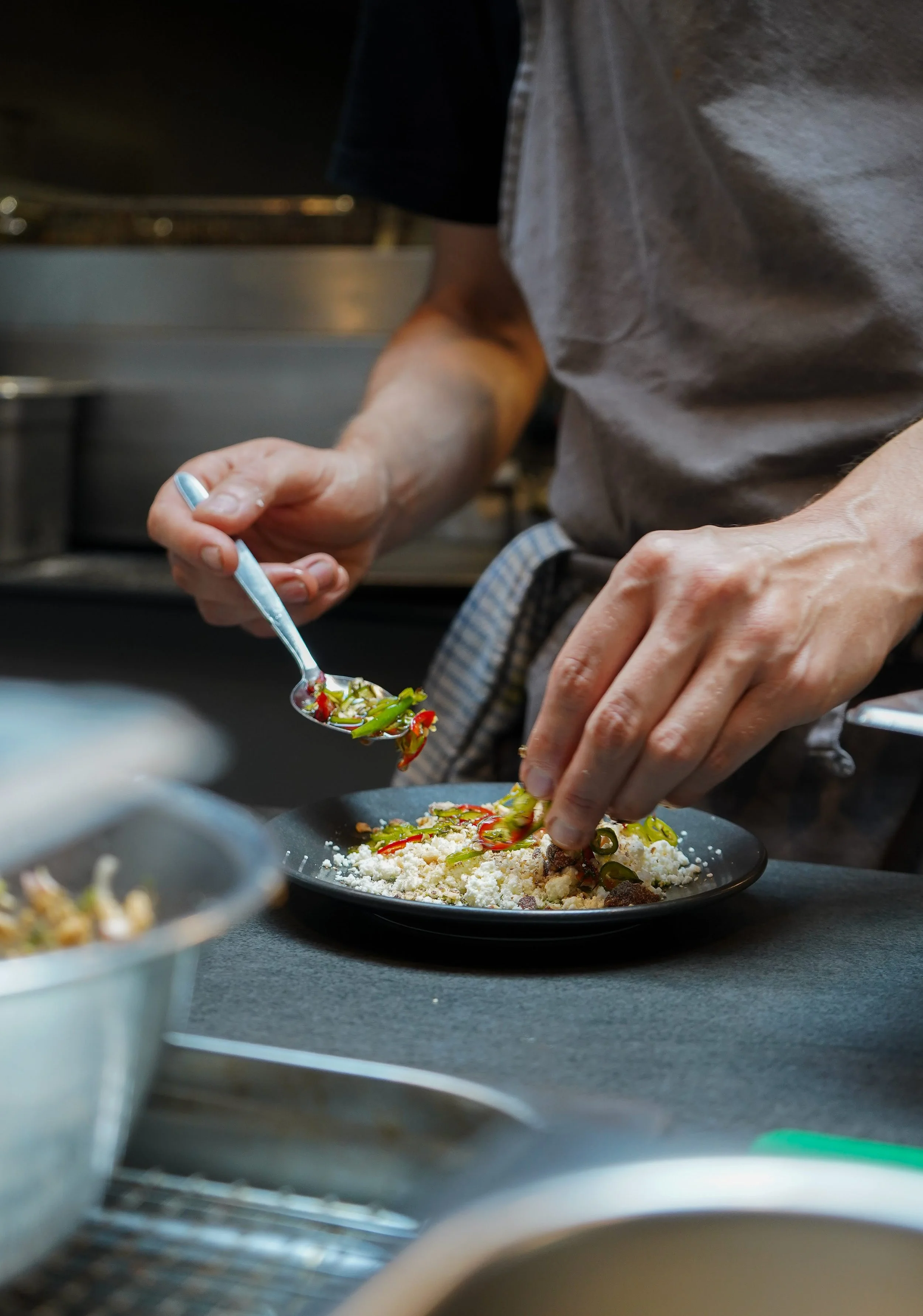 A chef plating a dish with vegetables, crumbled cheese, and other ingredients in a professional kitchen.