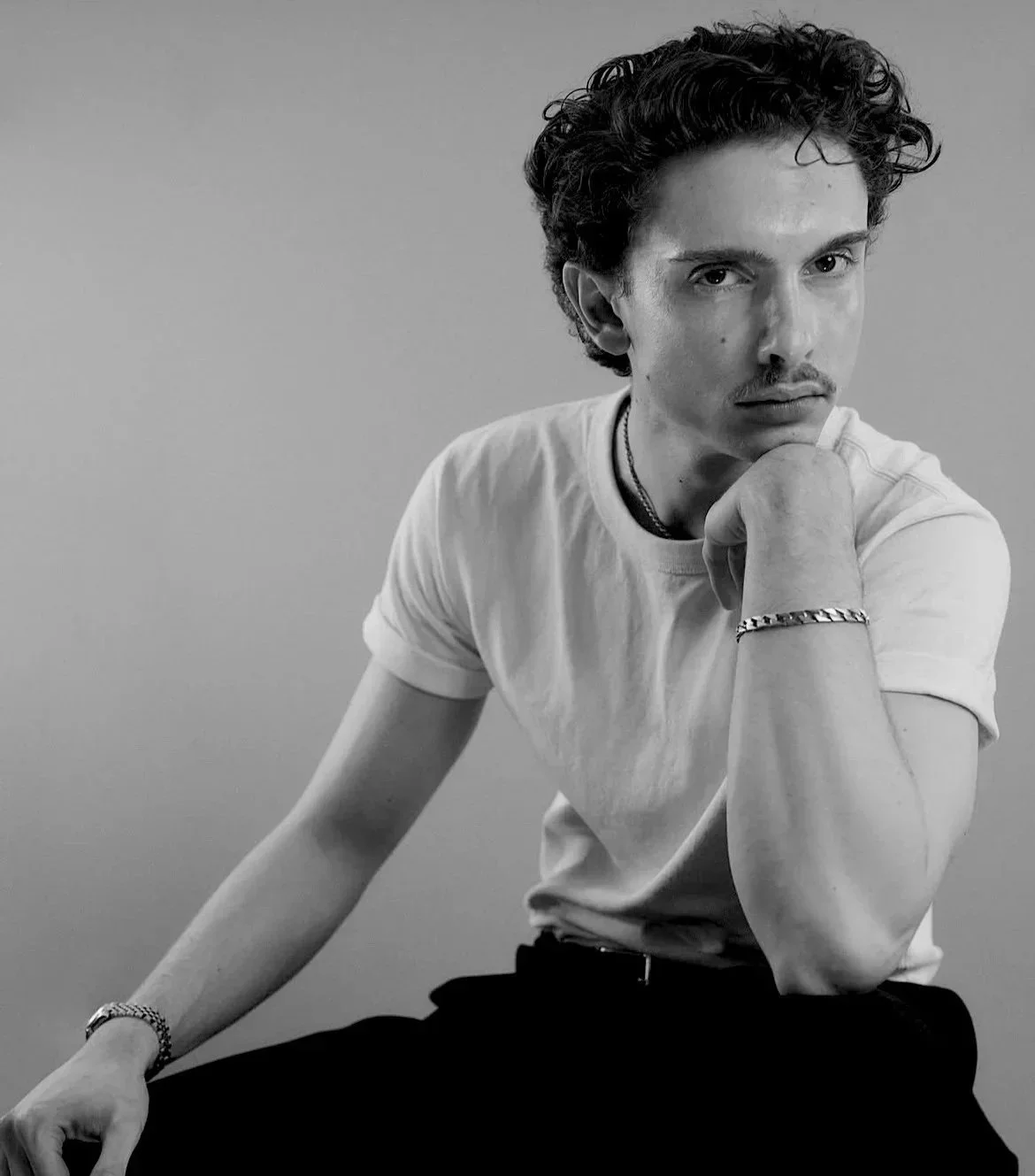 Black and white portrait of Agency PSG founder, Parker Gibson with short, curly hair, wearing a white t-shirt, sitting with one arm resting on his knee and his chin resting on his hand, looking thoughtfully into the camera.