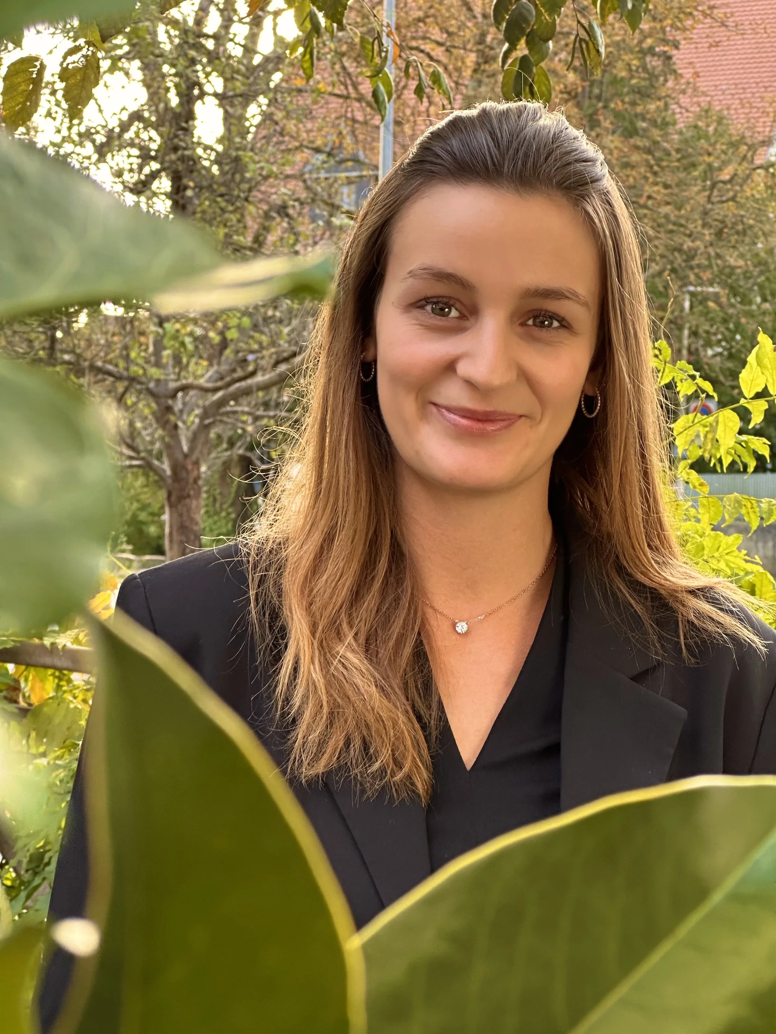Ein Lächeln tragende Frau mit braunen Haaren, die einen schwarzen Blazer trägt, steht draußen vor einem herbstlichen Baum mit roten Blättern, teilweise von großen grünen Blättern im Vordergrund verdeckt.