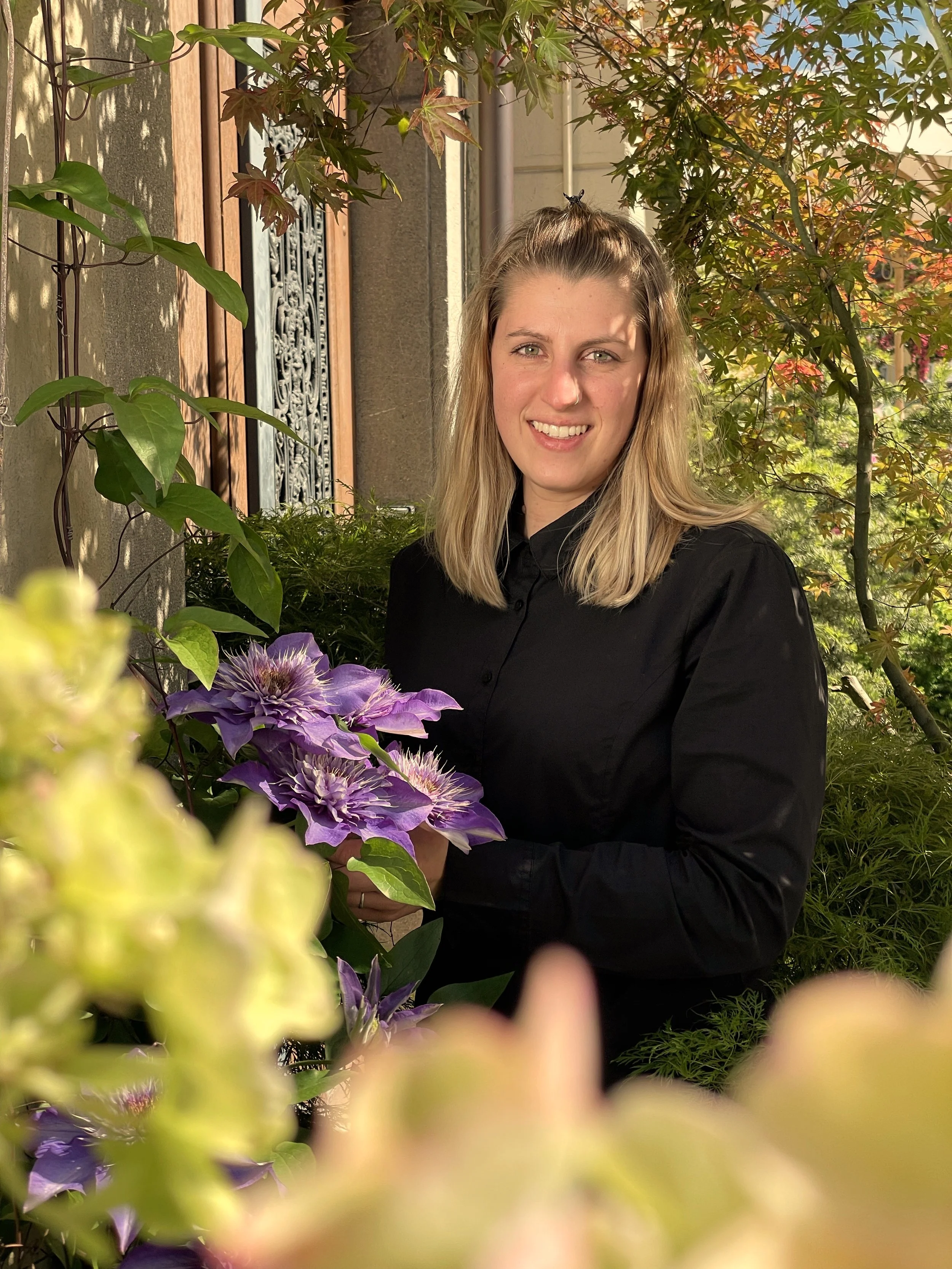 Junge Frau mit blonden Haaren, schwarzer Bluse, sitzt im Garten vor einem Baum mit roten und grünen Blättern, im Vordergrund bunte Blumen. Sonniger Tag.