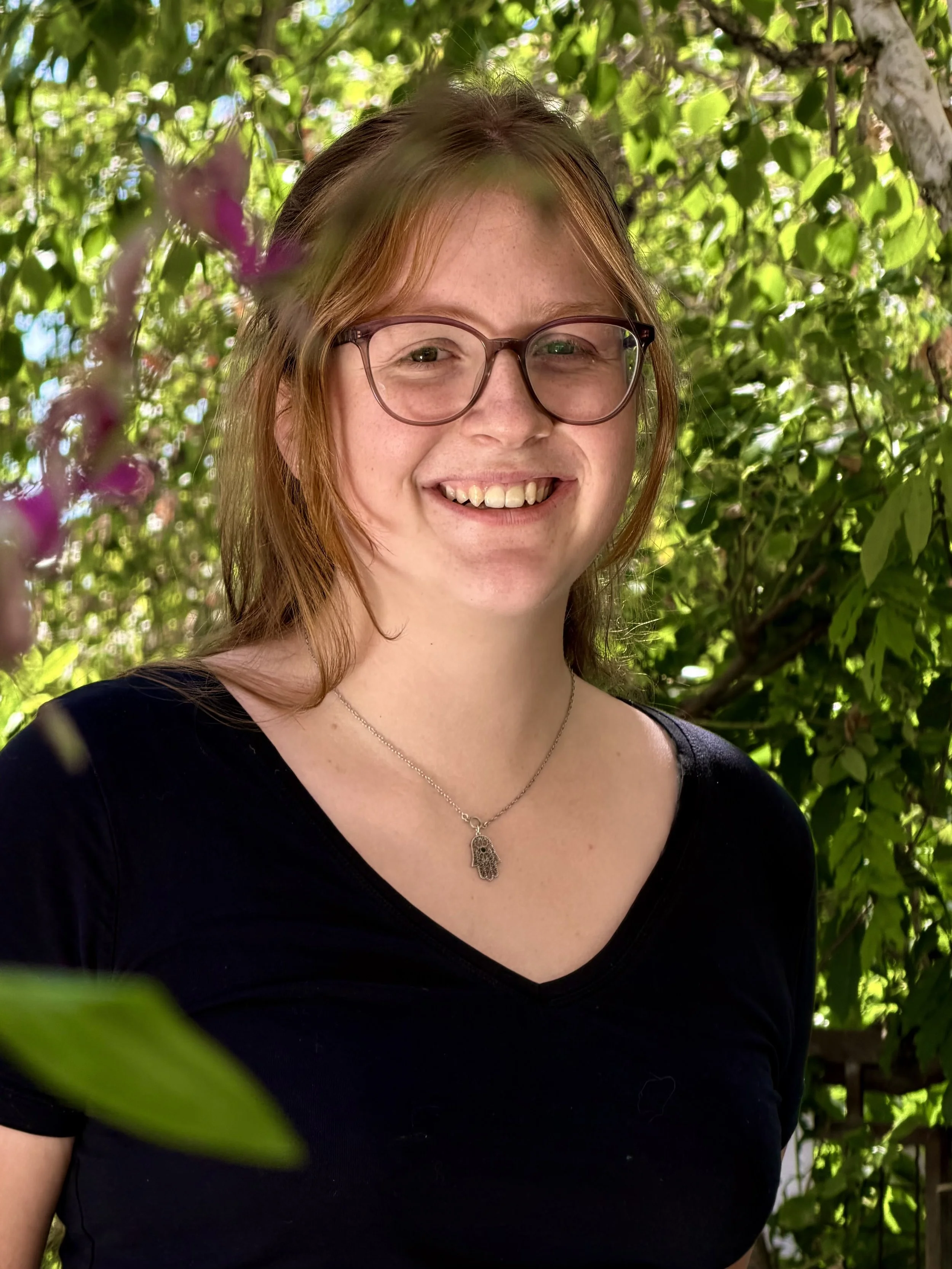 Ein lächelnder junger Frau mit Brille und Kette, im Freien vor einem grünen Baum.