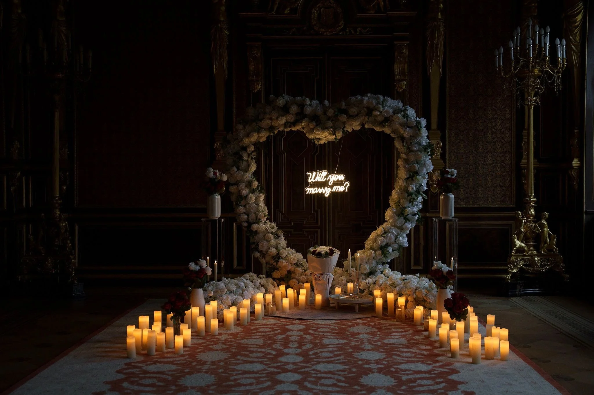 Romantische Verlobungsdekoration mit großem herzförmigen Blumenrahmen aus weißen Rosen, zahlreichen Kerzen am Boden und einem leuchtenden Schriftzug „Will you marry me?“ vor einer dunklen Wand.