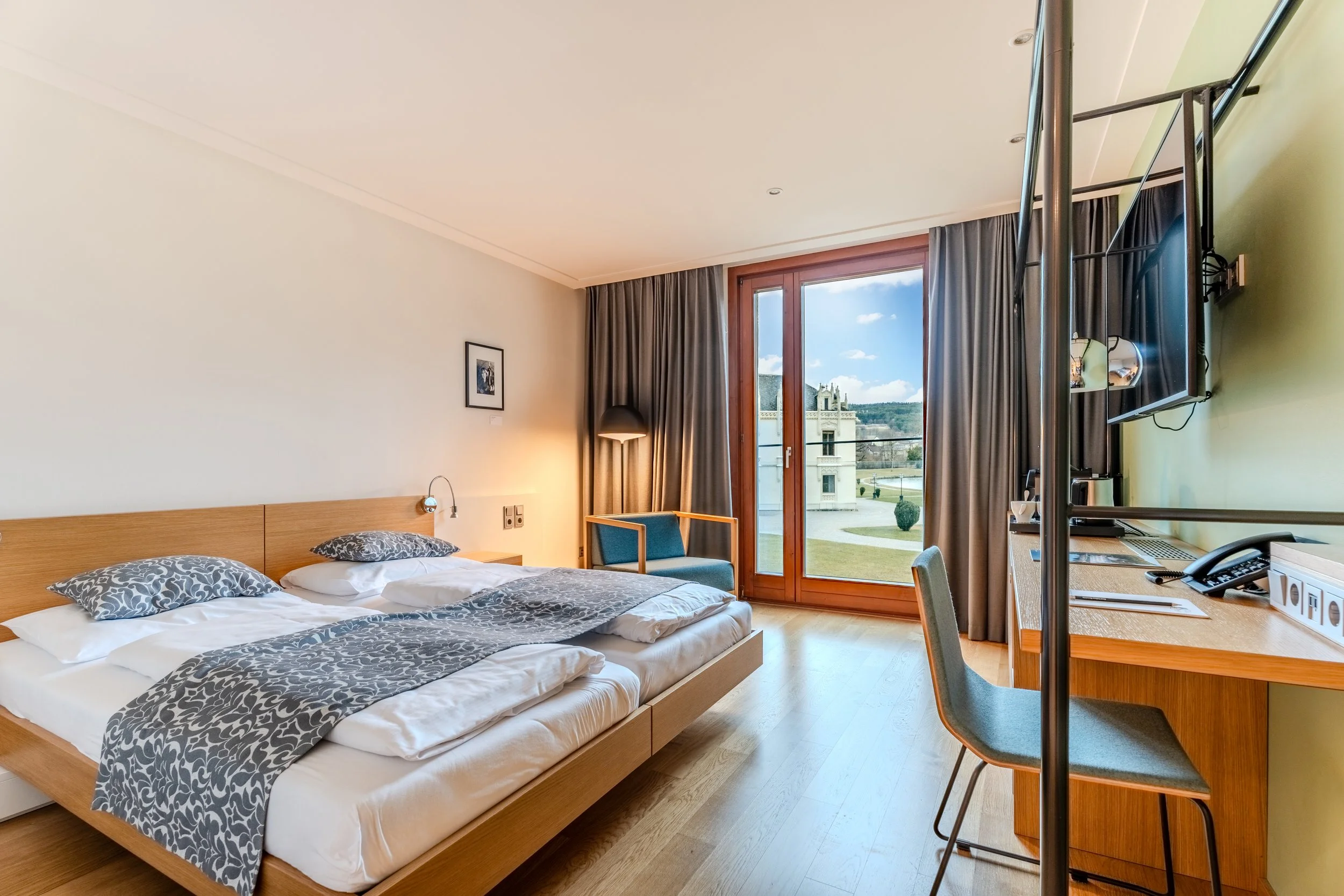 Helles Hotelzimmer mit Doppelbett, Holzmöbeln und großem Fenster mit Blick auf eine grüne Landschaft und ein Gebäude im Freien; daneben ein Schreibtisch, Stuhl und Vorhänge.