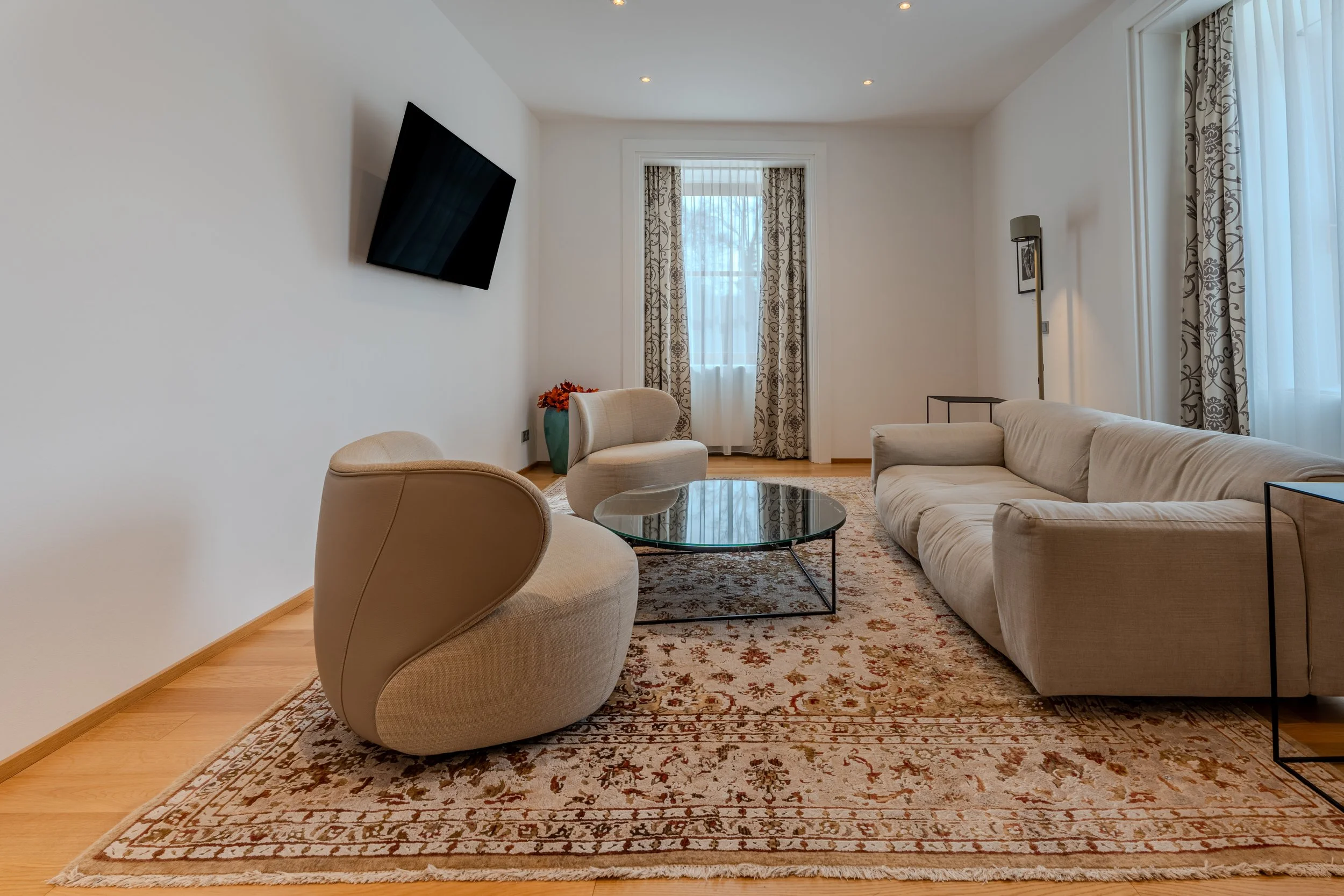 Modernes, helles Wohnzimmer mit großem Sofa, zwei bequemen Sesseln, rundem Glastisch und Teppich, wandmontiertem Fernseher sowie großen Fenstern mit Vorhängen und warmen Holzboden