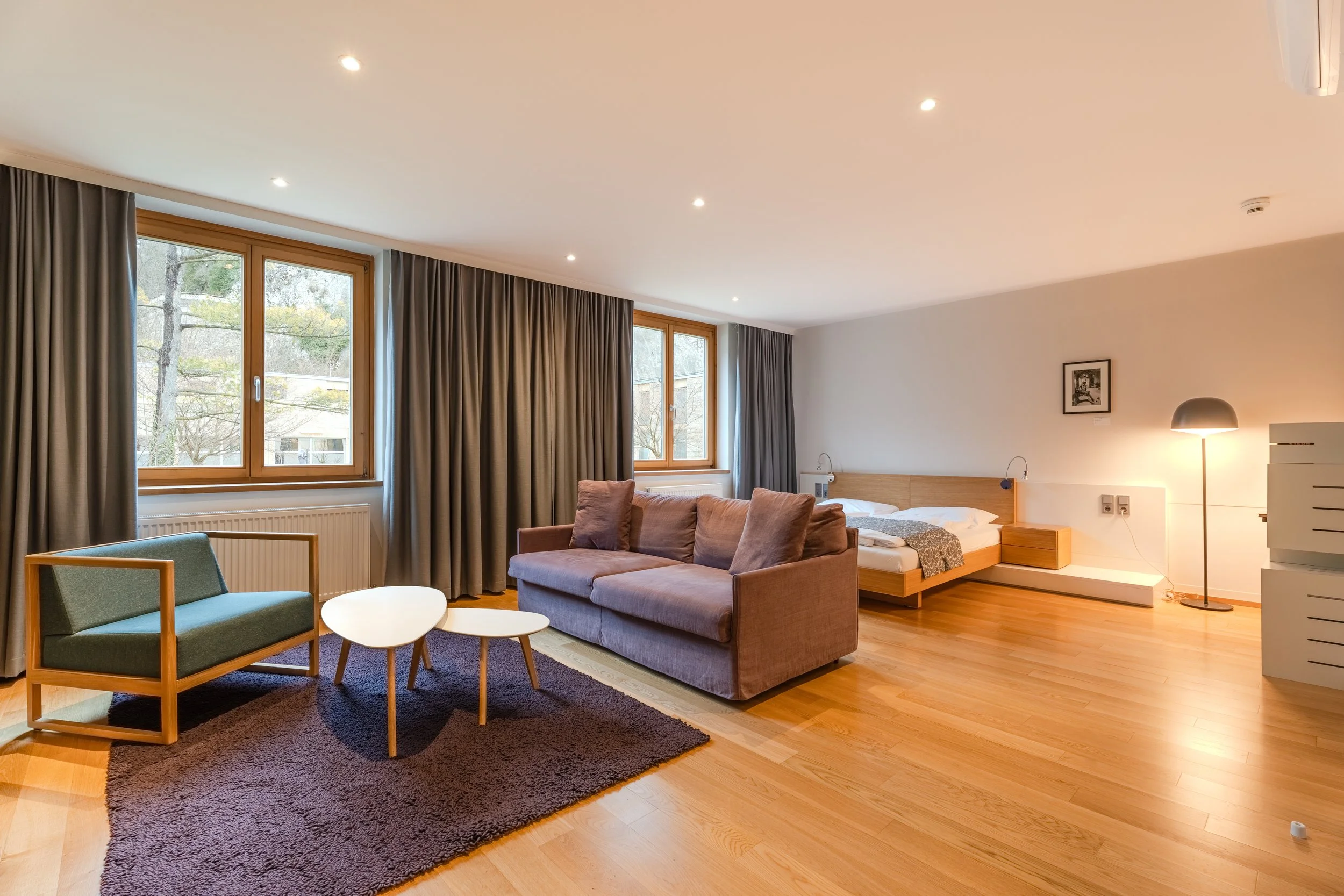 Heller Wohn- und Schlafbereich mit Sofa, Couchtischen, Teppich und Doppelbett nahe großer Fensterfront