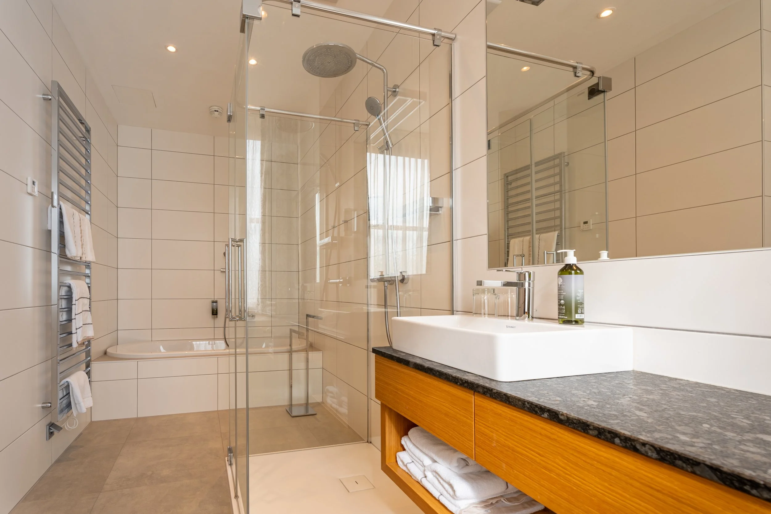 Modernes Badezimmer mit bodengleicher Glasdusche (Regendusche und Handbrause), Badewanne im Hintergrund, großem Waschbecken auf Holzunterschrank, Spiegel, Handtuchheizkörper und hellen Fliesen.