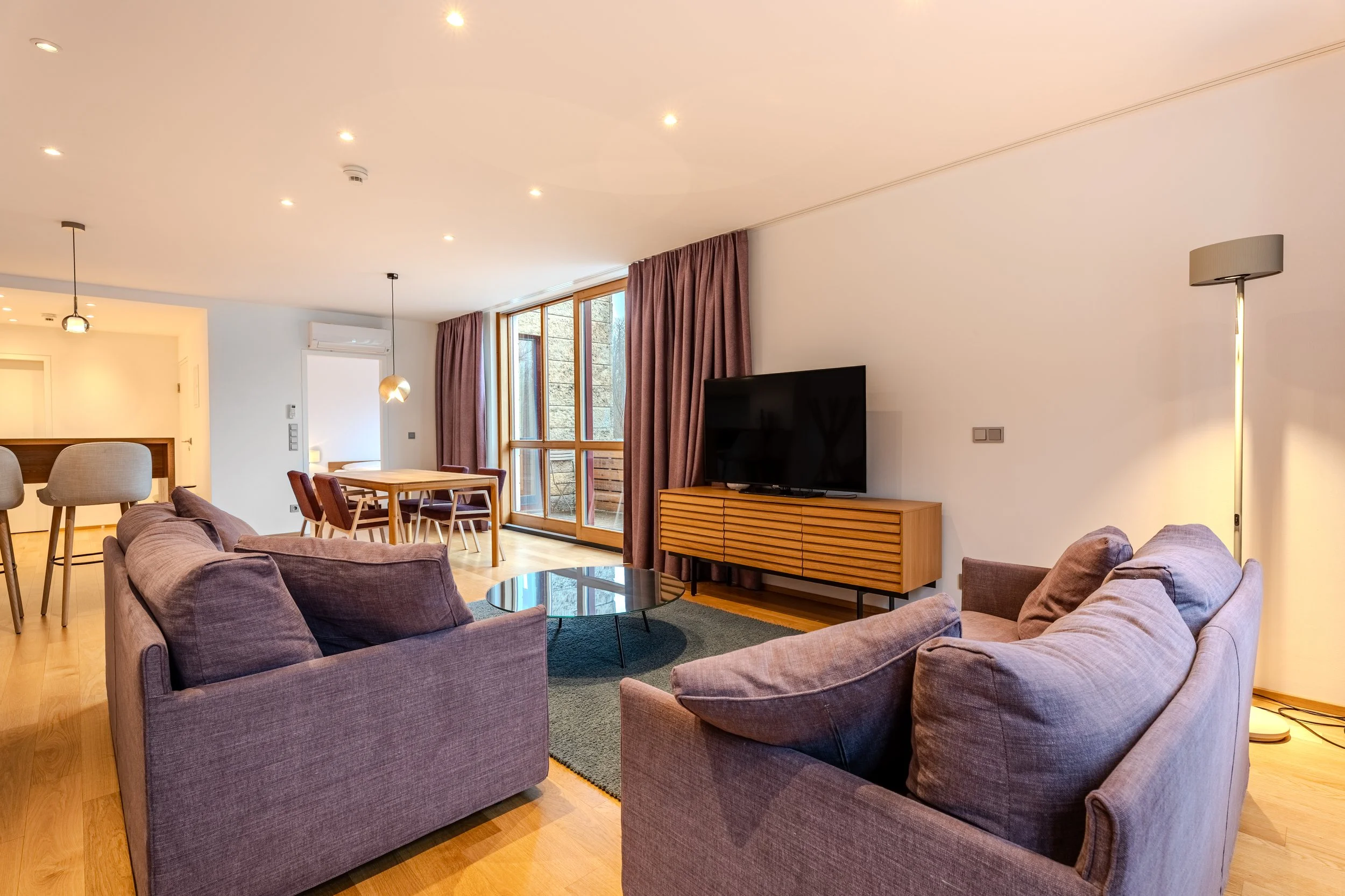 Gemütliches Wohnzimmer mit Sofa, Sessel, Couchtisch und Fernseher auf einem Lowboard, große Fenster mit Vorhängen.