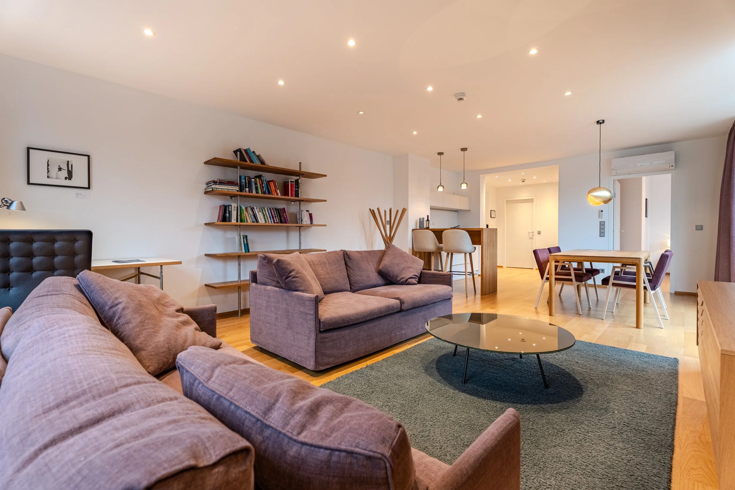 Moderner Wohnraum mit Sofa, rundem Couchtisch, Regalwand und offenem Essbereich mit Tisch und Stühlen.