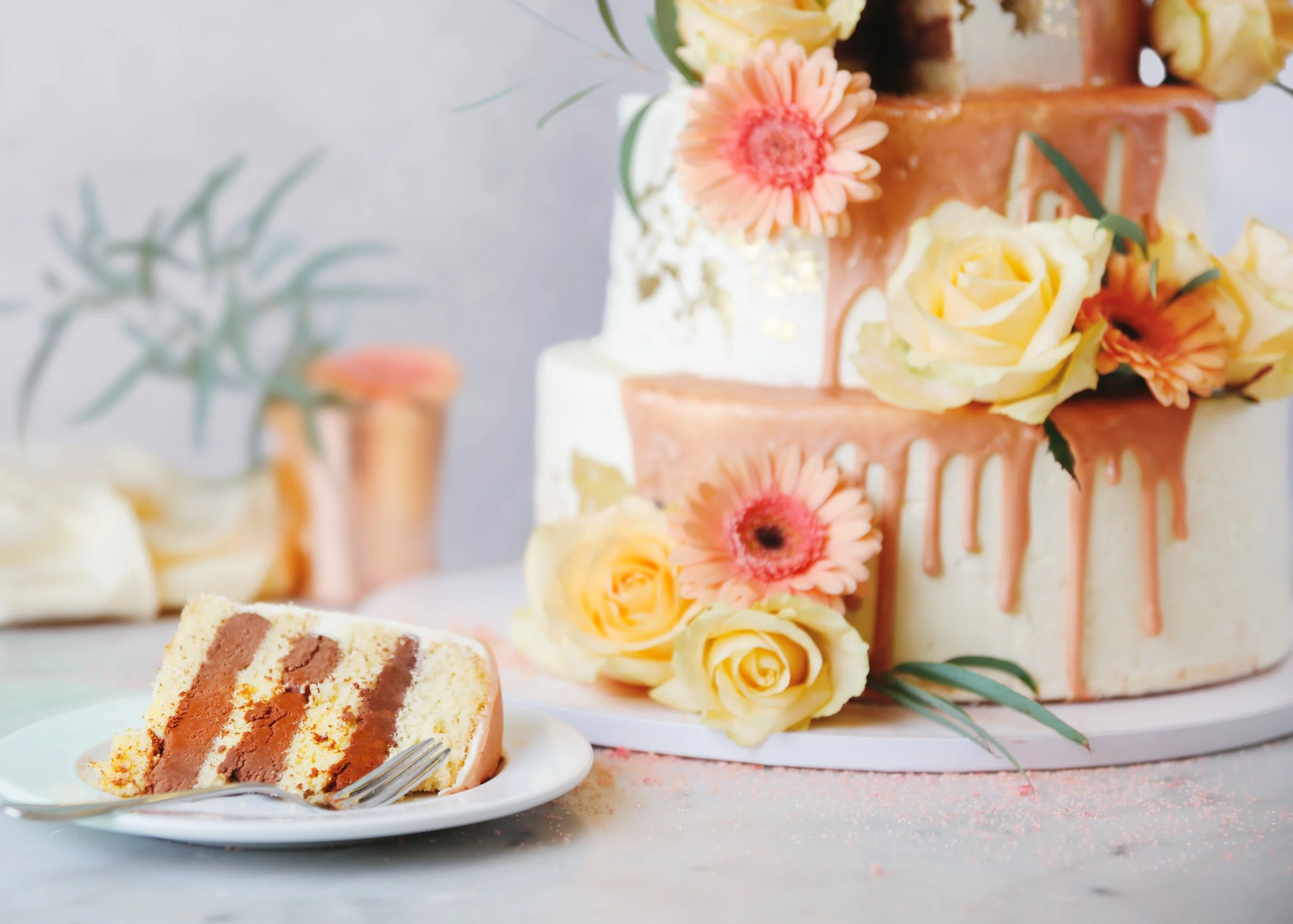 Mehrstöckige Hochzeitstorte mit roségoldenem Drip, dekoriert mit gelben Rosen und Gerbera, daneben ein angeschnittenes Kuchenstück auf einem Teller mit Gabel