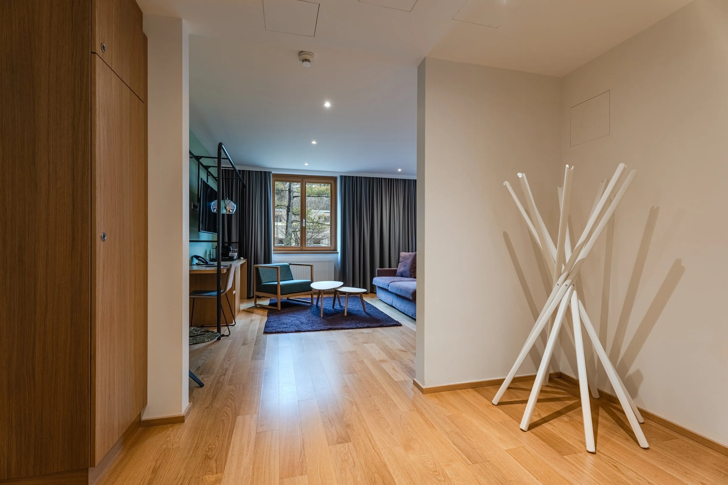 Flur mit Holzfußboden, Blick in einen Wohnbereich mit Sofa, Sessel, Schreibtisch und großen Fenstern.