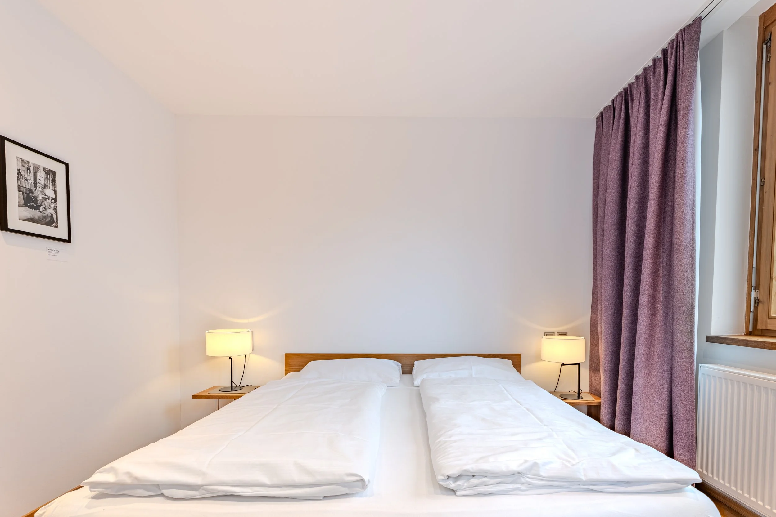 Schlafzimmer mit Doppelbett, Nachttischlampen, Holzboden und Fenster mit Vorhängen.