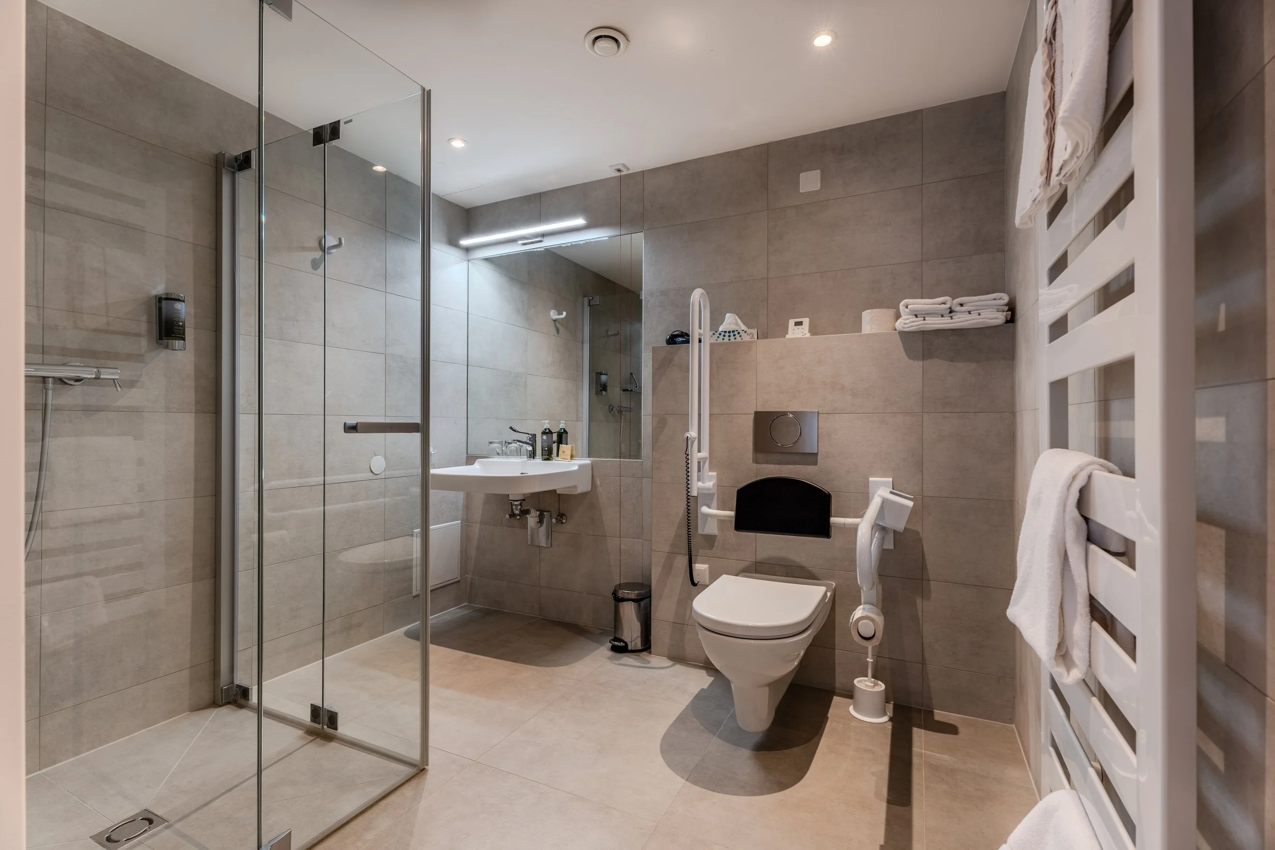 Modernes, barrierefreies Badezimmer mit bodengleicher Dusche aus Glas, wandhängendem WC mit Haltegriffen, unterfahrbarem Waschbecken und neutralen Fliesen in Beige- und Grautönen. Frische Handtücher liegen ordentlich bereit.