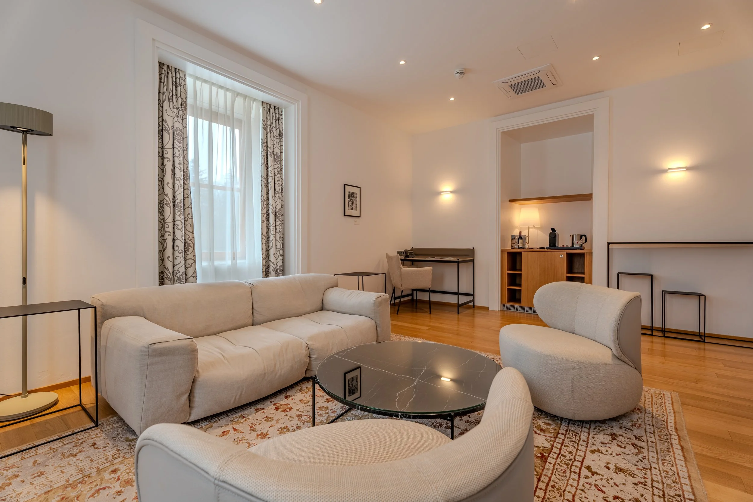 Gemütlicher Wohnbereich mit hellem Sofa, Sesseln, Couchtisch und kleinem Arbeits- oder Küchenbereich im Hintergrund