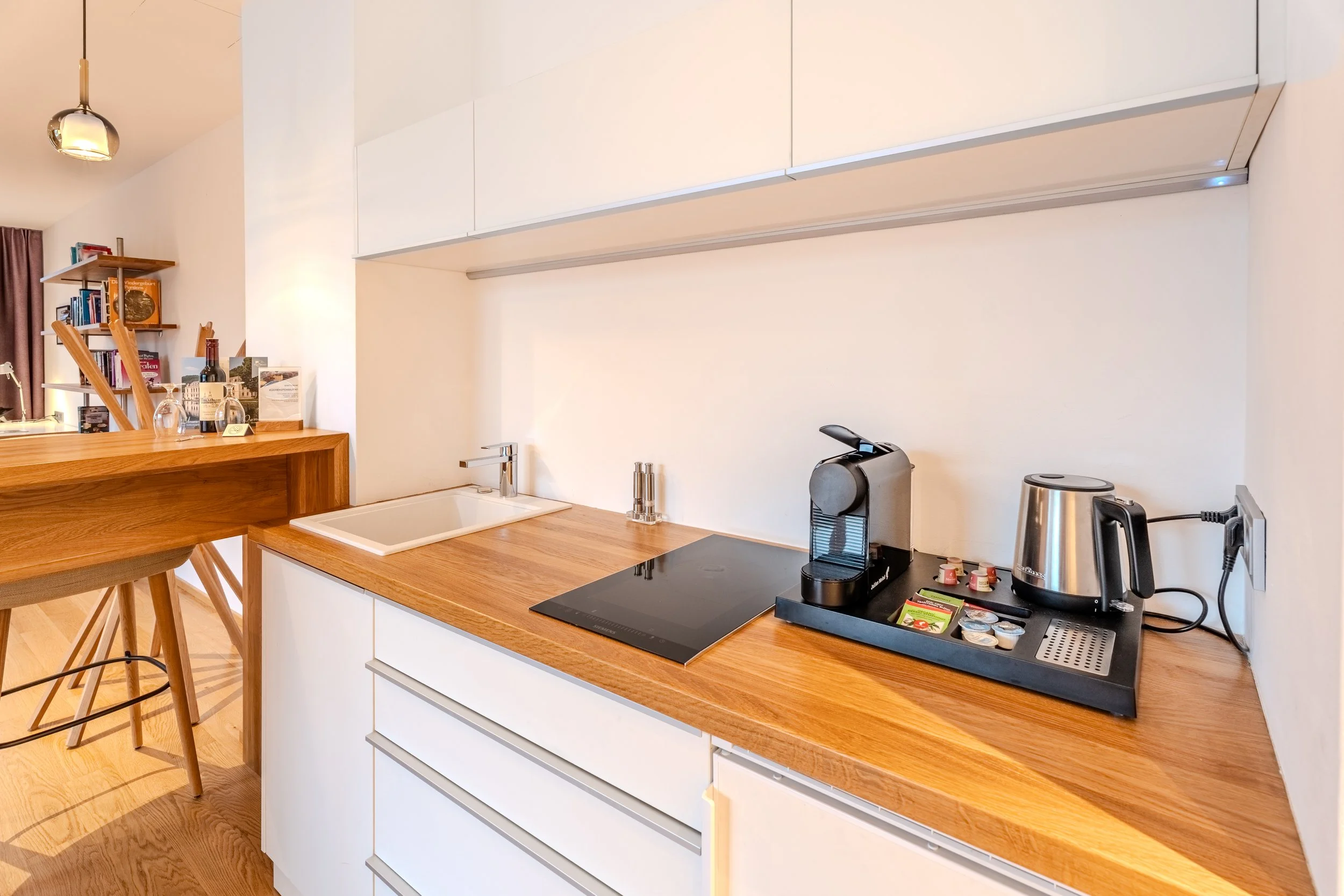 Moderne Küchenzeile mit Holzarbeitsplatte, integriertem Kochfeld, Kaffeemaschine, Wasserkocher und kleiner Bar mit Hockern.