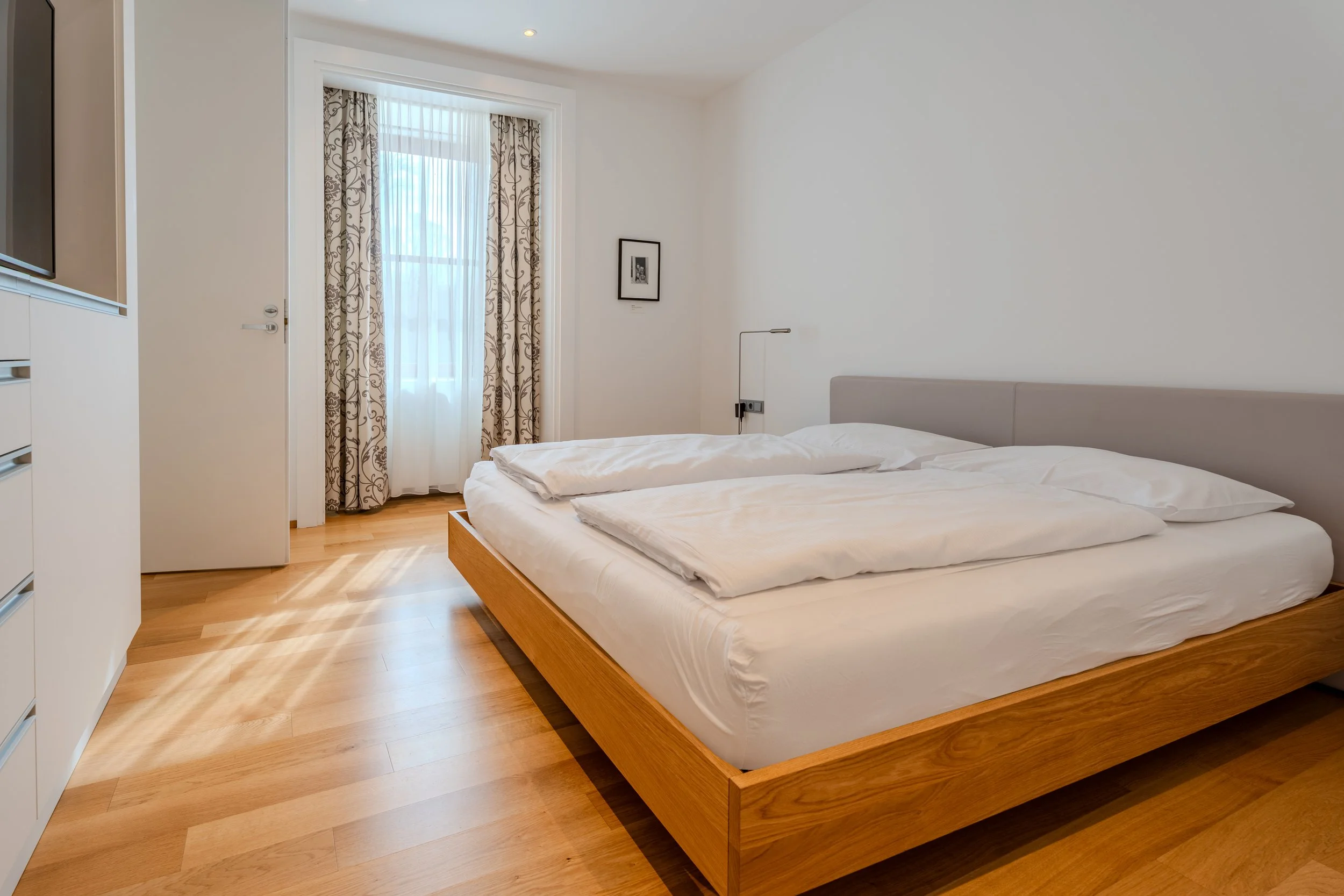 Schlafzimmer mit großem Doppelbett, Holzboden, Fenster mit Vorhängen und schlichter Einrichtung.