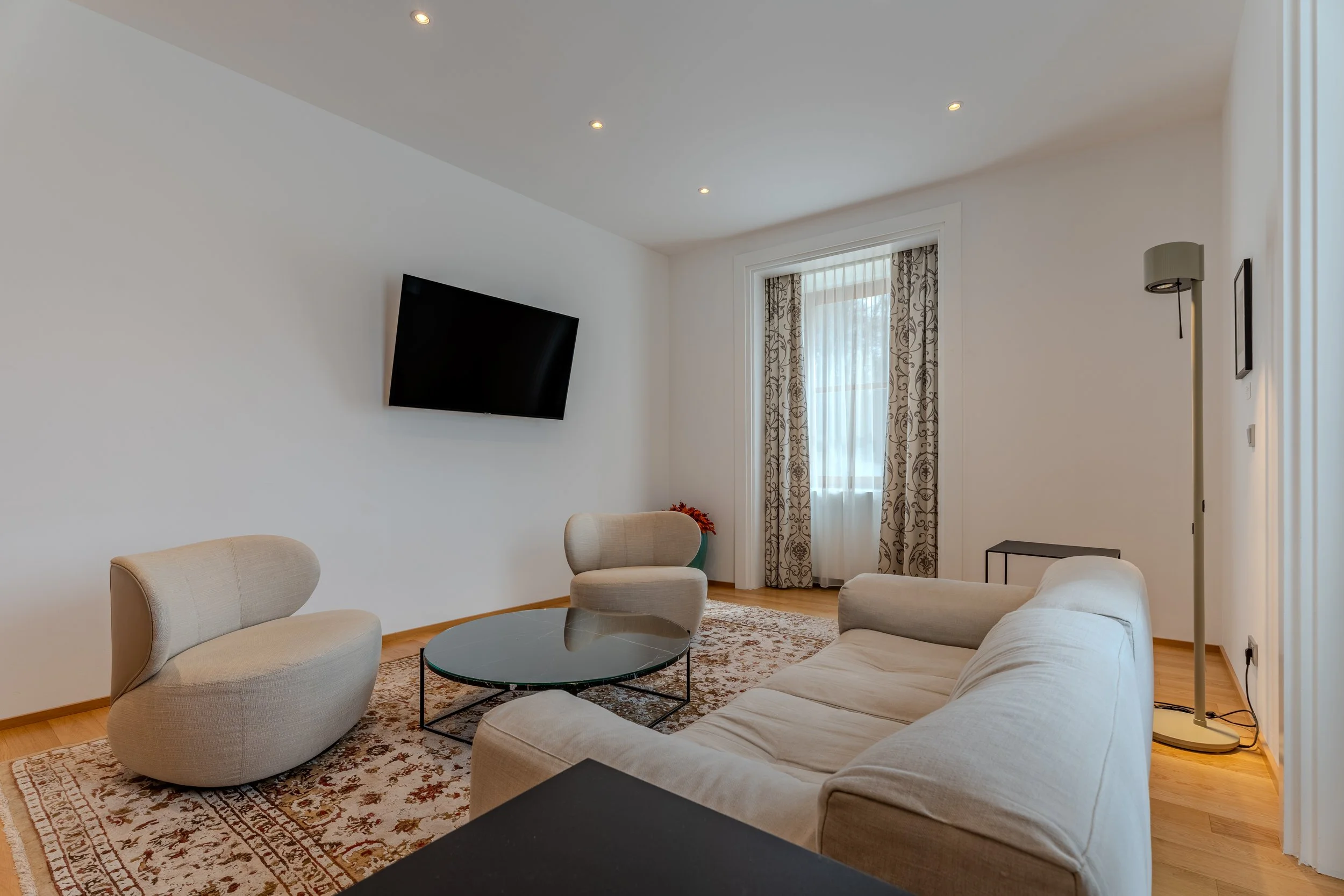 Wohnzimmer mit Sofa, Sesseln, Couchtisch, Teppich und wandmontiertem Fernseher in ruhiger Farbgestaltung.