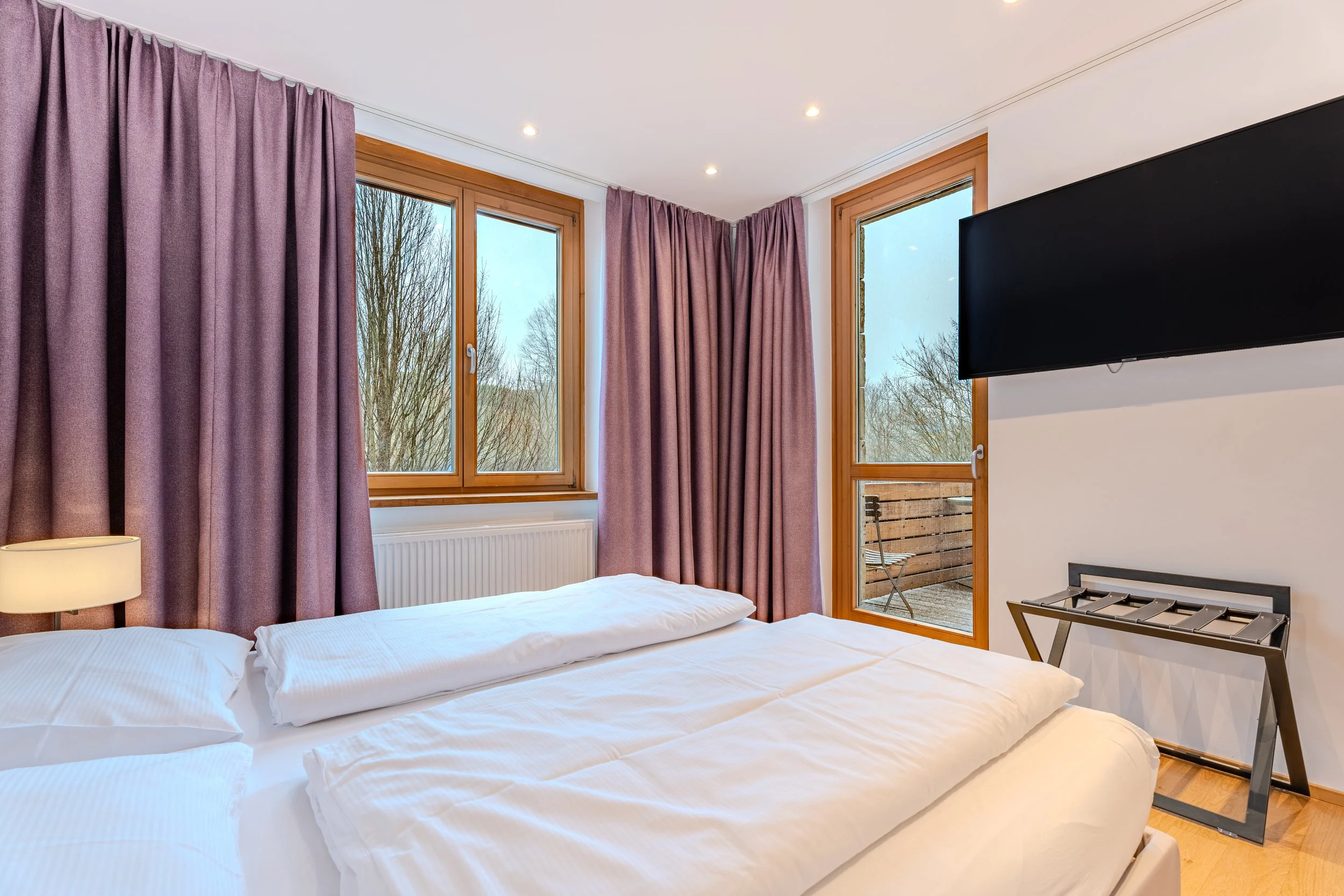 Helles Schlafzimmer mit Doppelbett, großen Fenstern mit Vorhängen, Wand-TV sowie Zugang zu einem Balkon.