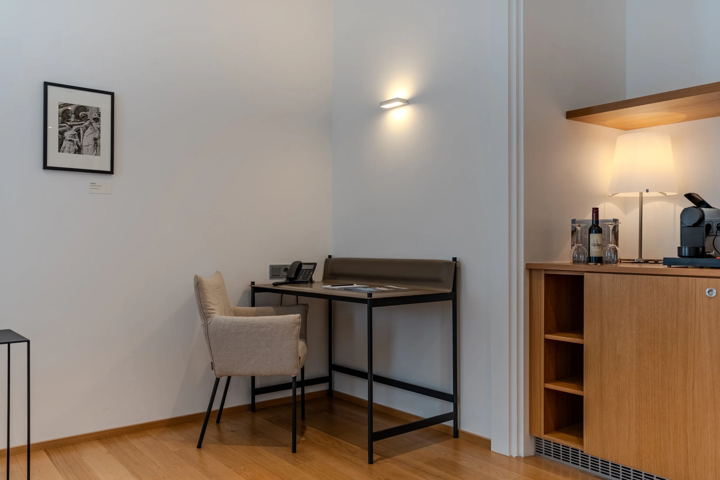 Kleiner Arbeitsbereich mit Schreibtisch, Stuhl, Telefon und Wandleuchte in minimalistisch gestaltetem Raum.