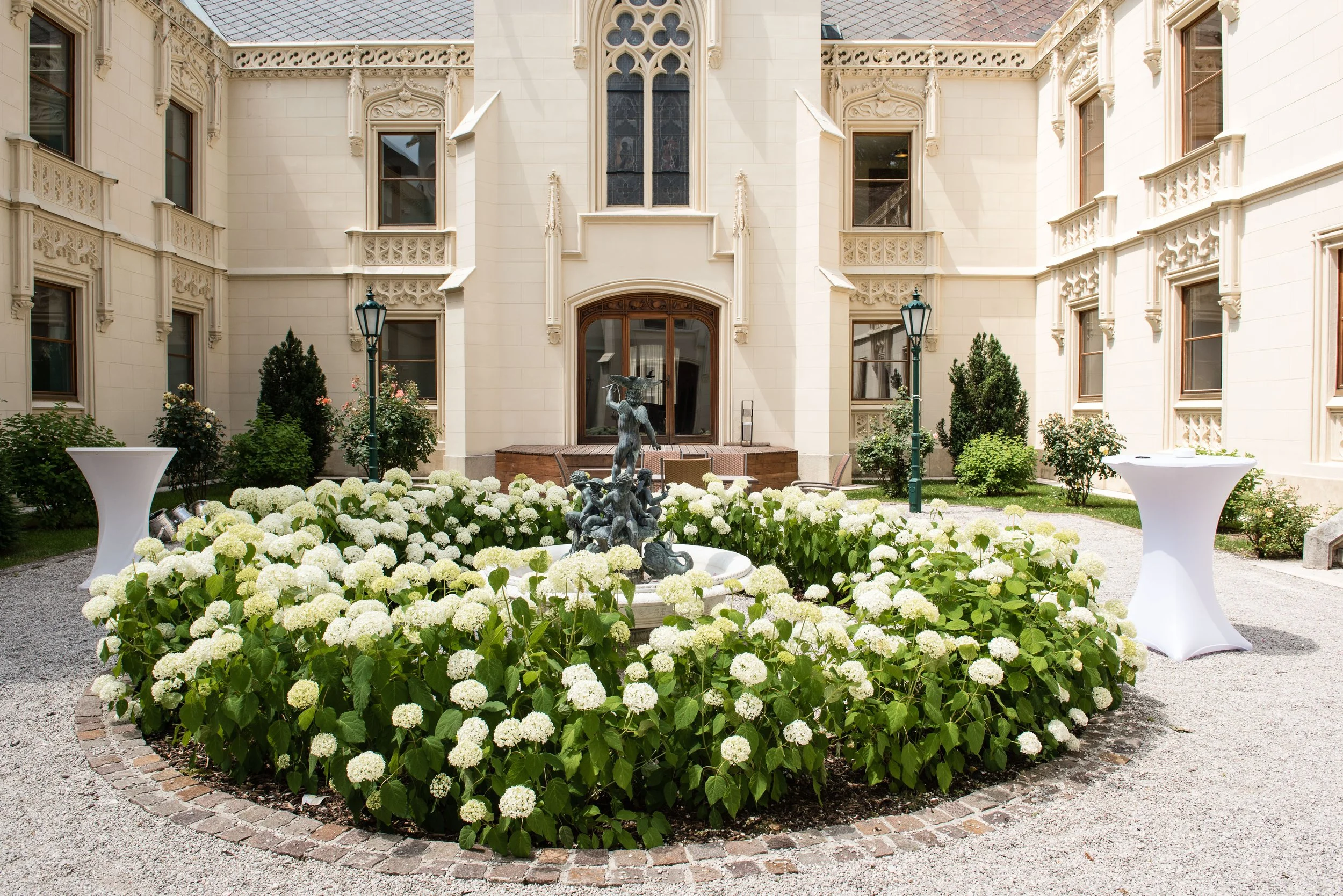 Eleganter Innenhof des Schloss Hernstein mit heller Fassade, verzierten Fenstern und einem zentralen Brunnen, umgeben von blühenden weißen Hortensien.