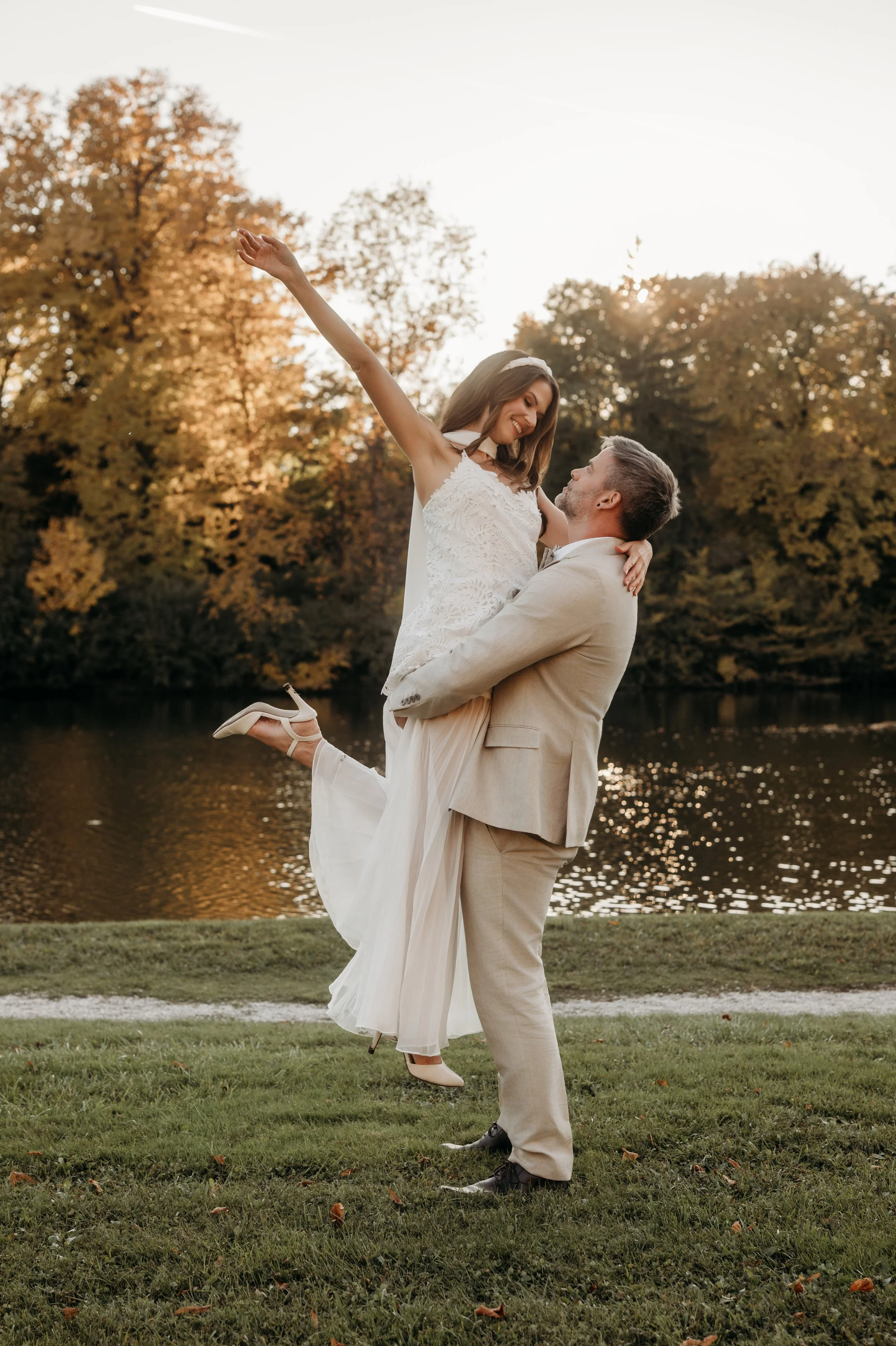Ein Paar im eleganter Hochzeitskleidung : Der Bräutigam hebt die Braut lachend hoch, während sie den Arm ausstreckt. Im Hintergrund ein ruhiger See und herbstlich gefärbte Bäume, die eine romantische Atmosphäre schaffen.