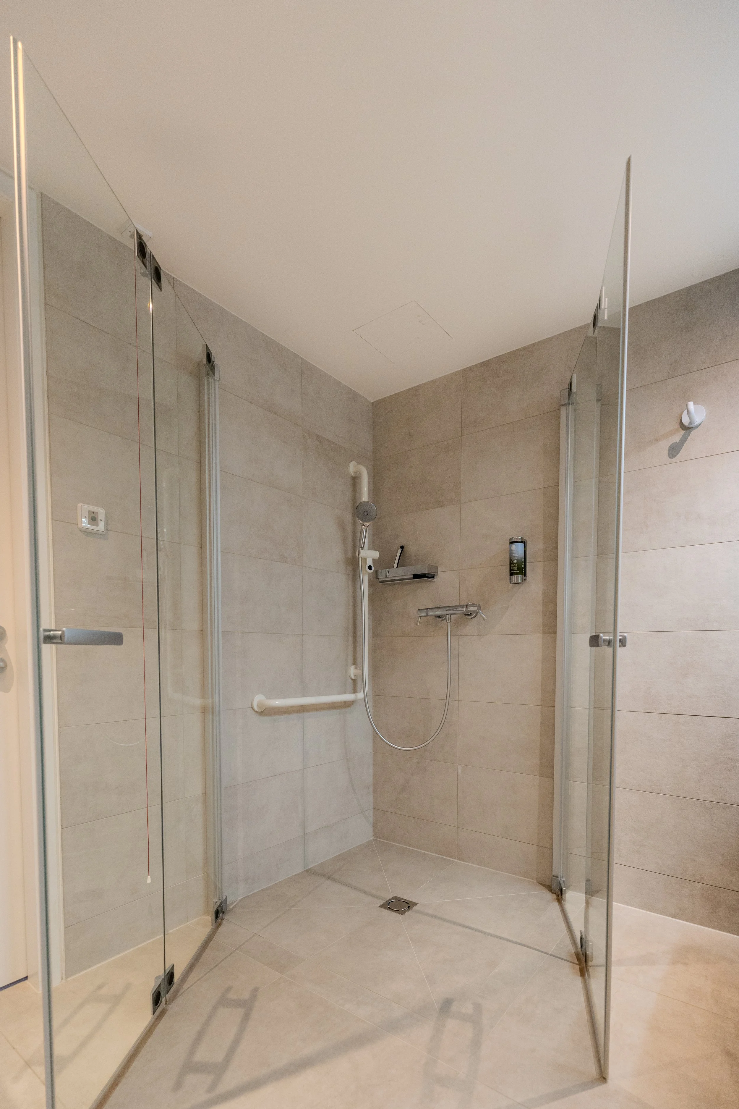 Barrierearmes Badezimmer mit bodengleicher Dusche, Haltegriffen und wandmontierter Duscharmatur in hellen Fliesen.