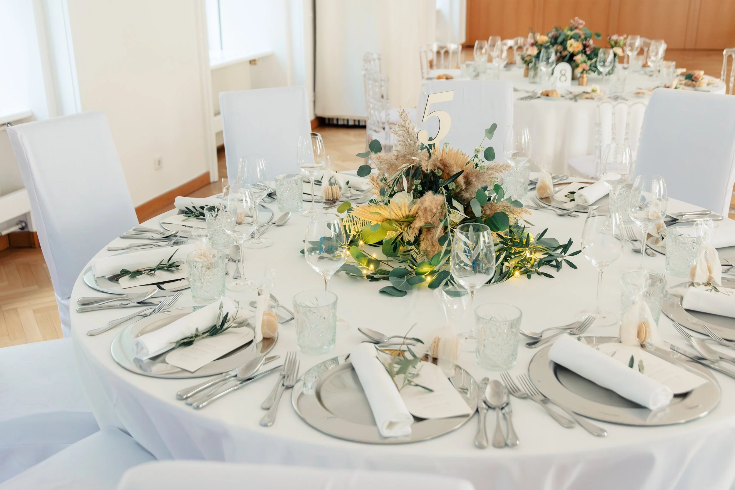 Festlich gedeckter runder Tisch mit Gläsern, Besteck und einer dekorativen Blumenarrangement-Mitte aus Trockenblumen und Grün, beleuchtet mit kleinen Lichtern – ideal für eine Feier oder Hochzeit.