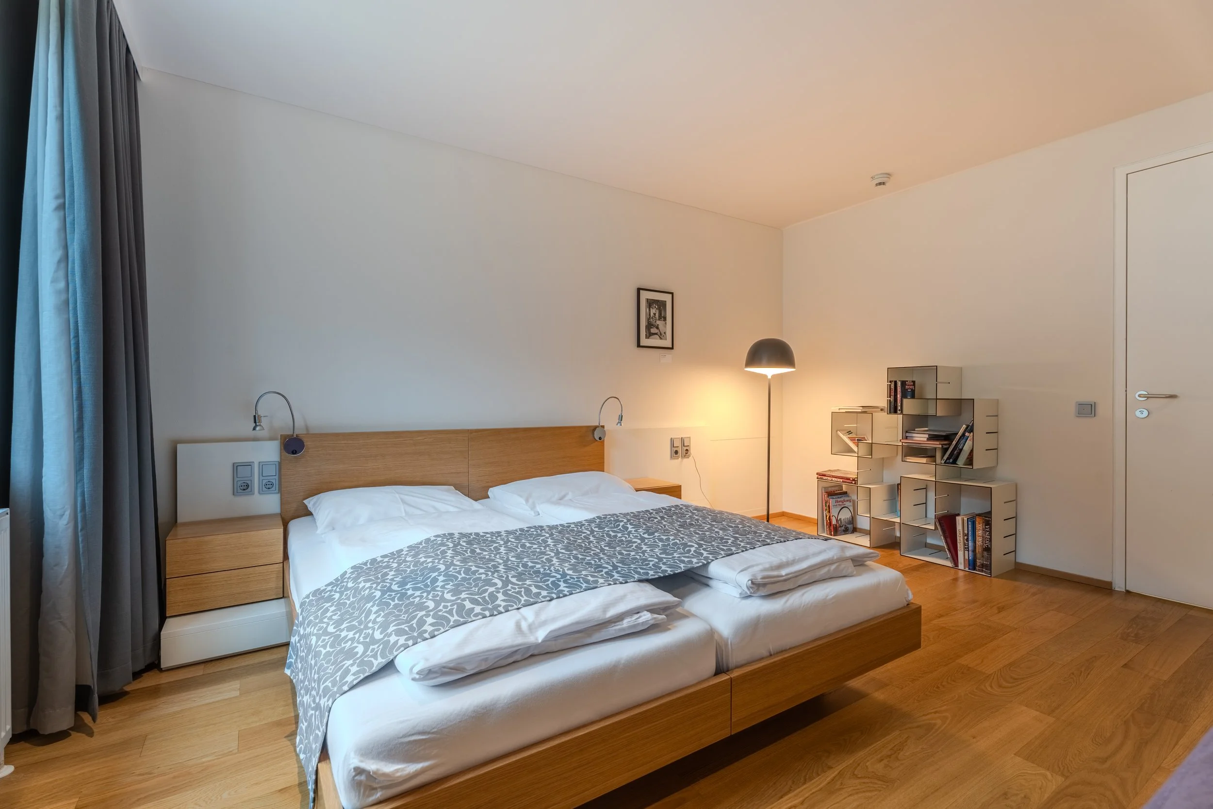 Helles, modernes Schlafzimmer mit Doppelbett, Holzrahmen und weißer Bettwäsche, dekorativer Tagesdecke, zwei Leselampen am Kopfteil, Nachttischen sowie einem offenen Regal und Stehlampe auf warmem Holzboden.