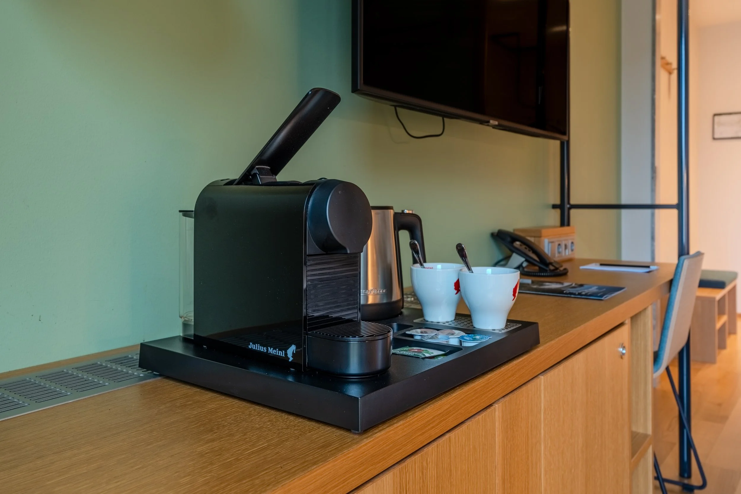 Kaffeemaschine auf einem Holzschreibtisch mit Tassen, Wasserkocher und Zubehör in einem Hotelzimmer.